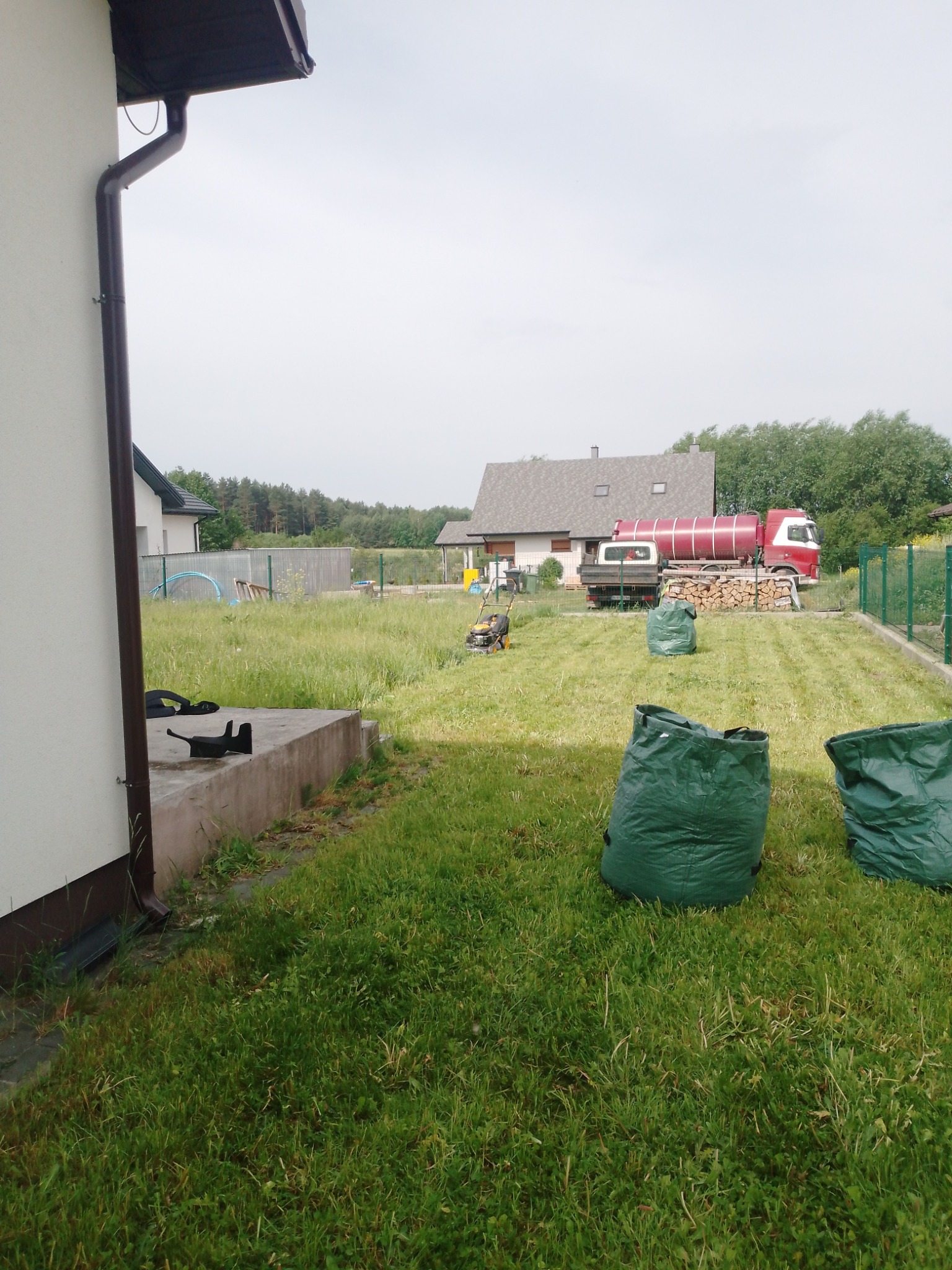 Trawnik po częściowym skoszeniu, widoczne trzy wypełnione worki na trawę, kosiarka w tle oraz ciężarówka z cysterną.