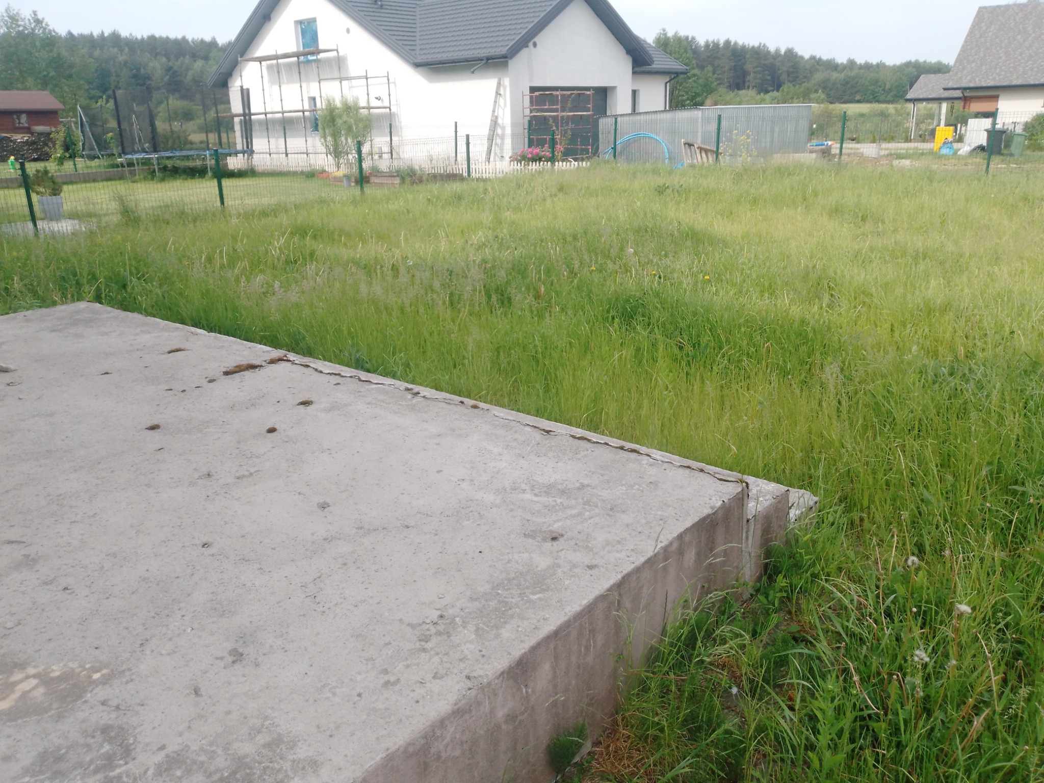 Betonowa podstawa w trawie przed domem z rusztowaniem i wysoką trawą.