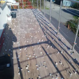 Układanie drewnopodobnych płytek na balkonie, widoczne krzyżyki dystansowe, narzędzia pracy i cień osoby fotografującej.