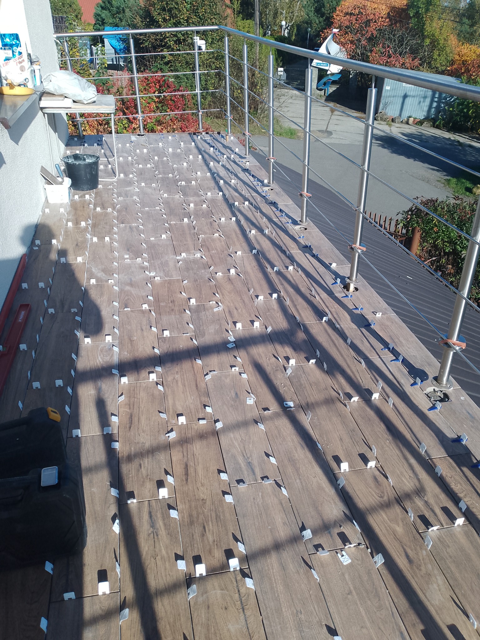 Układanie drewnopodobnych płytek na balkonie, widoczne krzyżyki dystansowe, narzędzia pracy i cień osoby fotografującej.