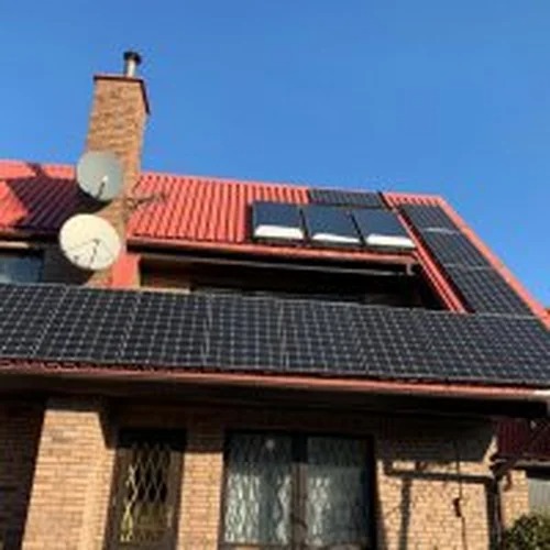 Ceglany dom jednorodzinny z czerwonym dachem, na którym zamontowano panele fotowoltaiczne i kolektory słoneczne; widoczne dwie anteny satelitarne na kominie.