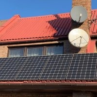 Panele słoneczne na dachu domu z czerwoną dachówką, widoczne dwie anteny satelitarne na kominie. Ujęcie z dołu, lekko z prawej strony.
