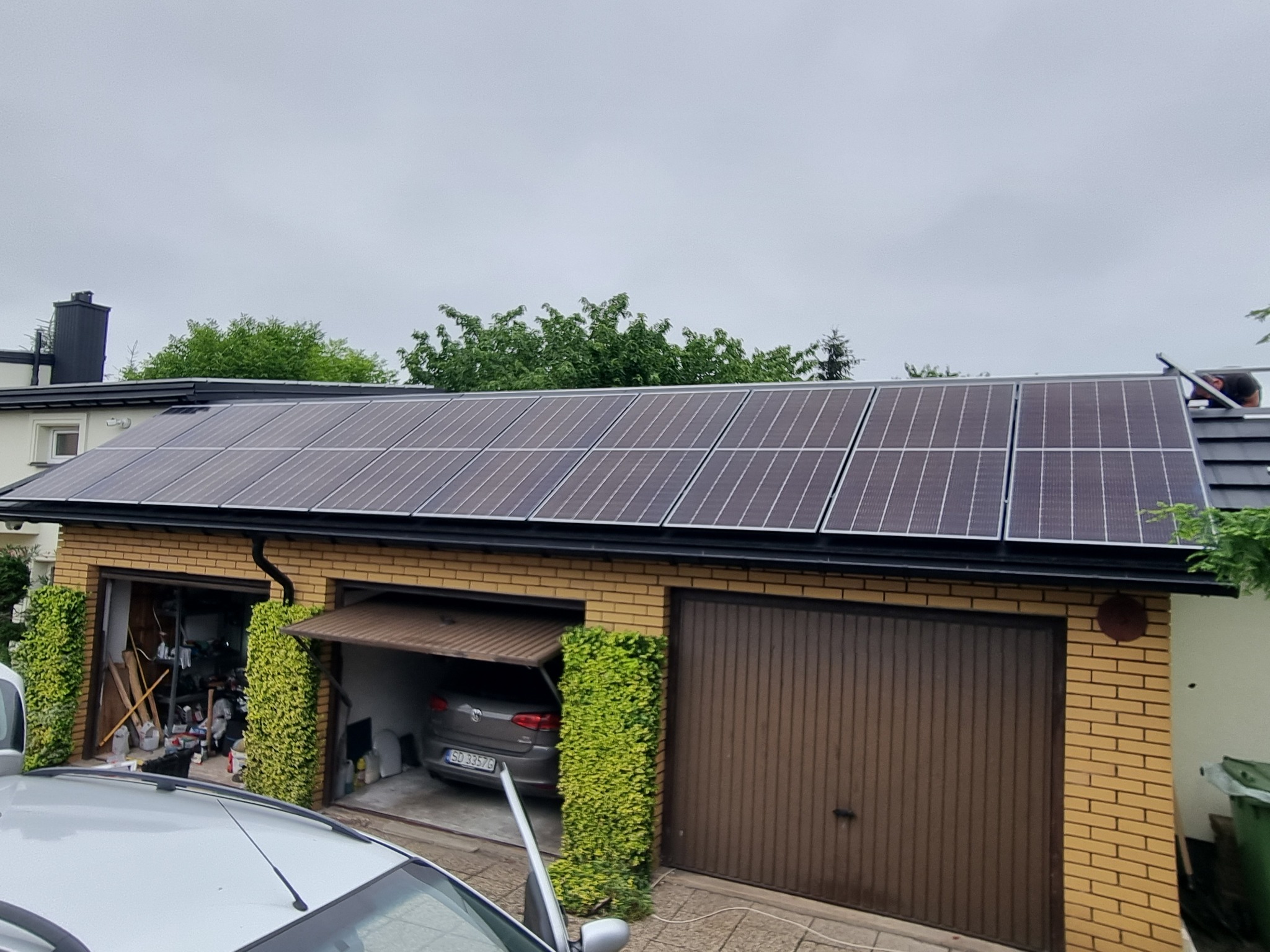 Panele słoneczne na dachu garażu z cegły, częściowo przysłoniętego przez roślinność, z samochodem w otwartym garażu i pochmurnym niebem w tle.