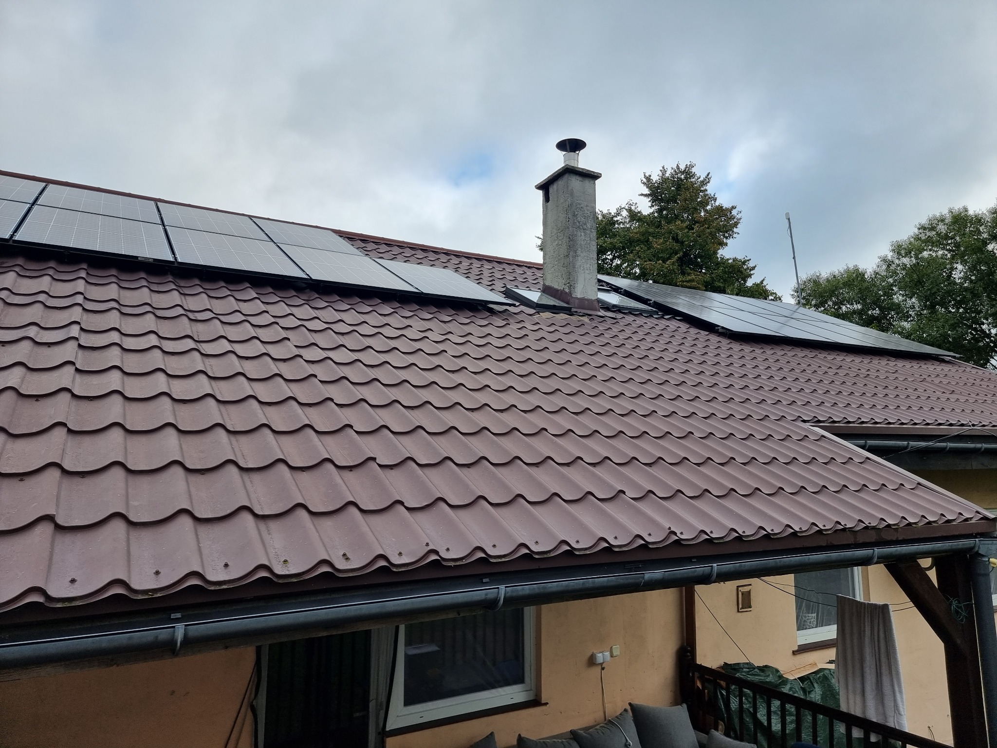 Brązowy dach z panelami słonecznymi i betonowym kominem na tle pochmurnego nieba i drzew.