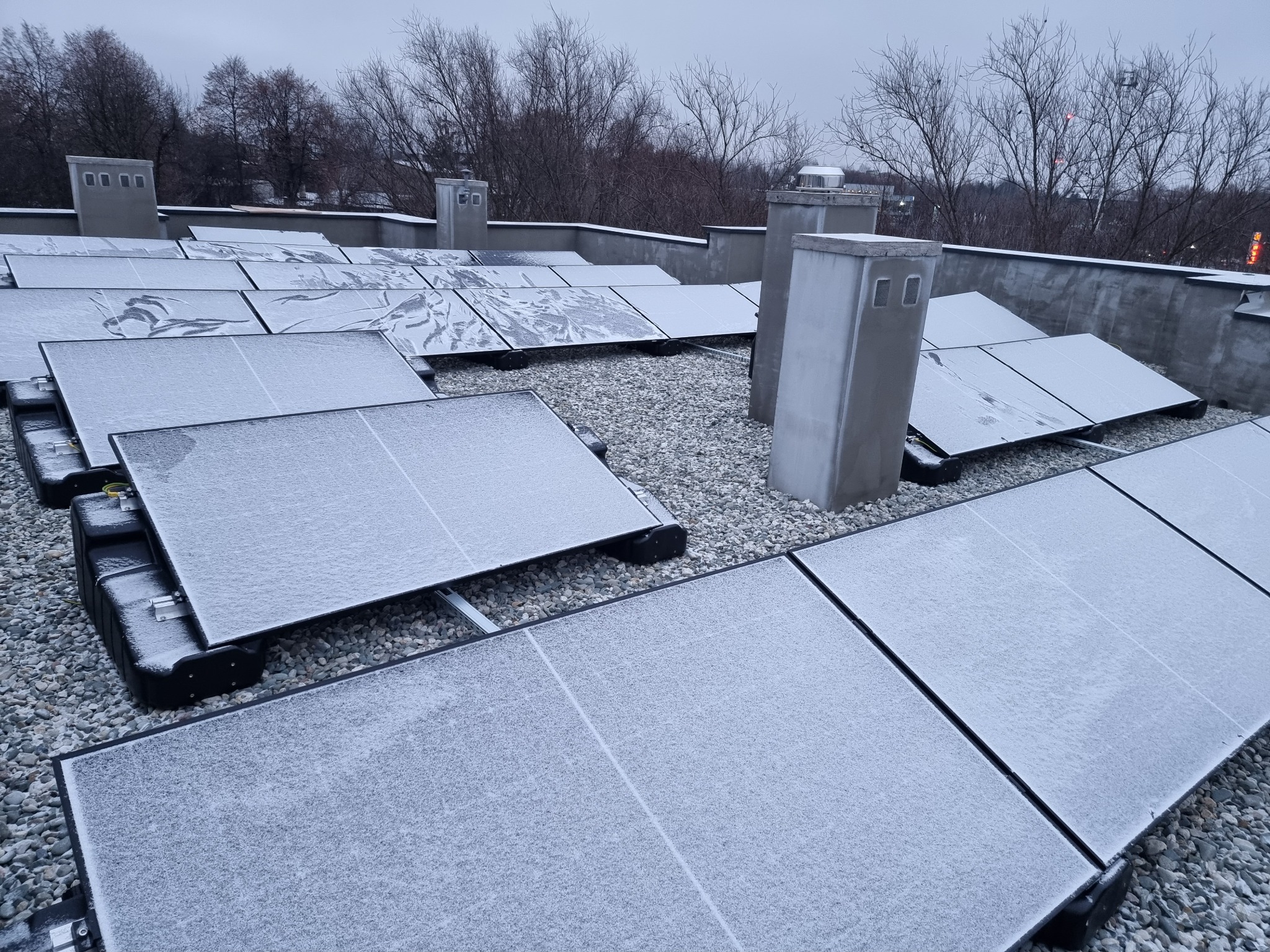 Instalacja fotowoltaiczna na dachu pokryta szronem, widoczne panele, szare kamyki, kominy wentylacyjne i drzewa w tle, pochmurny dzień.