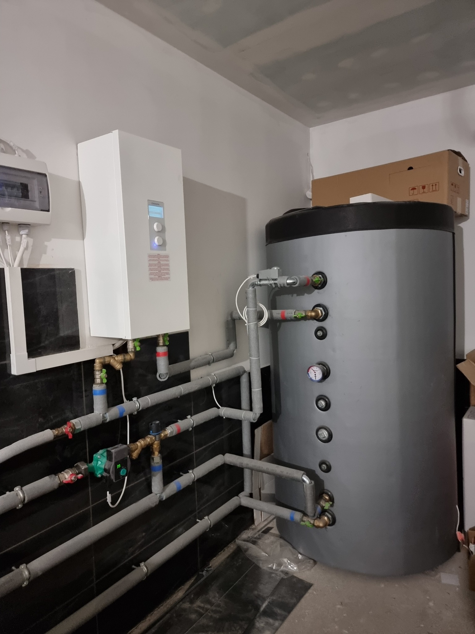 Instalacja centralnego ogrzewania z białym piecem i szarym zasobnikiem wody, widoczne rury z izolacją, manometr i skrzynka elektryczna na ścianie, w tle kartony.