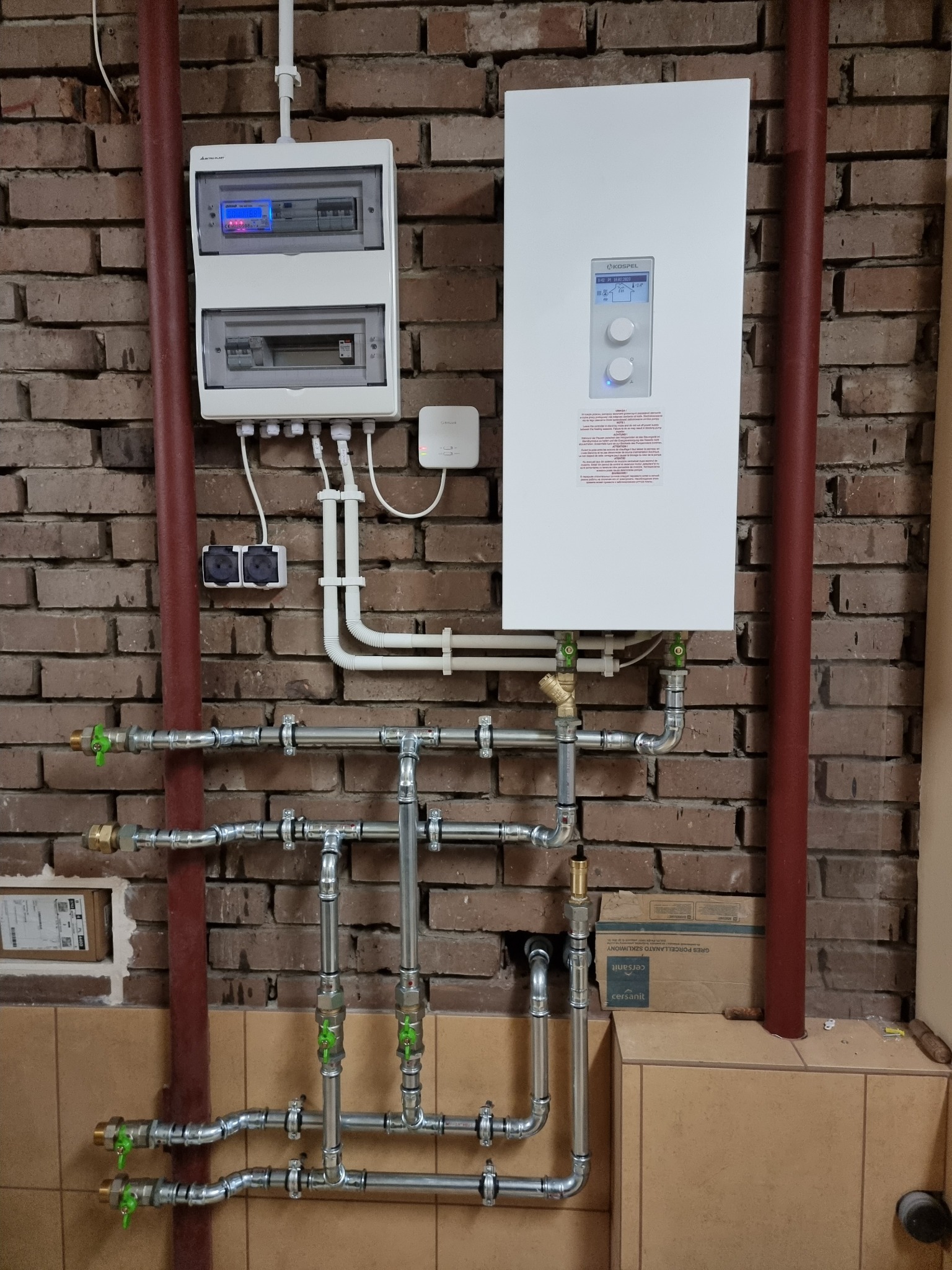 Biały panel sterowania i piec grzewczy na ceglanej ścianie, widoczne rury instalacji grzewczej z zielonymi zaworami, oraz skrzynka elektryczna z licznikiem.