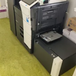 Wielofunkcyjne urządzenie biurowe Konica Minolta Bizhub C759 z otwartym podajnikiem papieru, widoczne zapasy papieru i tonera na półce w tle.