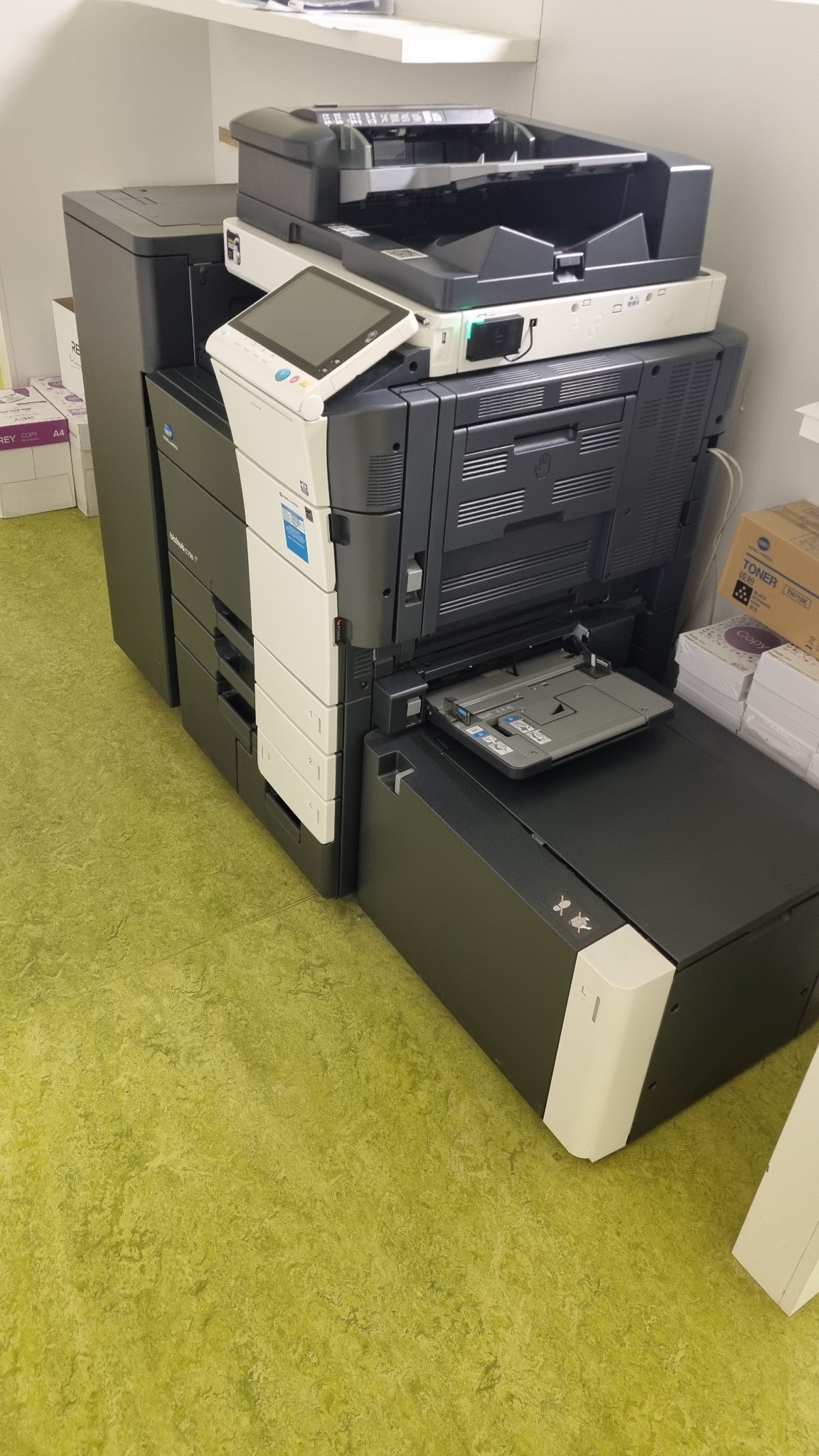 Wielofunkcyjne urządzenie biurowe Konica Minolta Bizhub C759 z otwartym podajnikiem papieru, widoczne zapasy papieru i tonera na półce w tle.