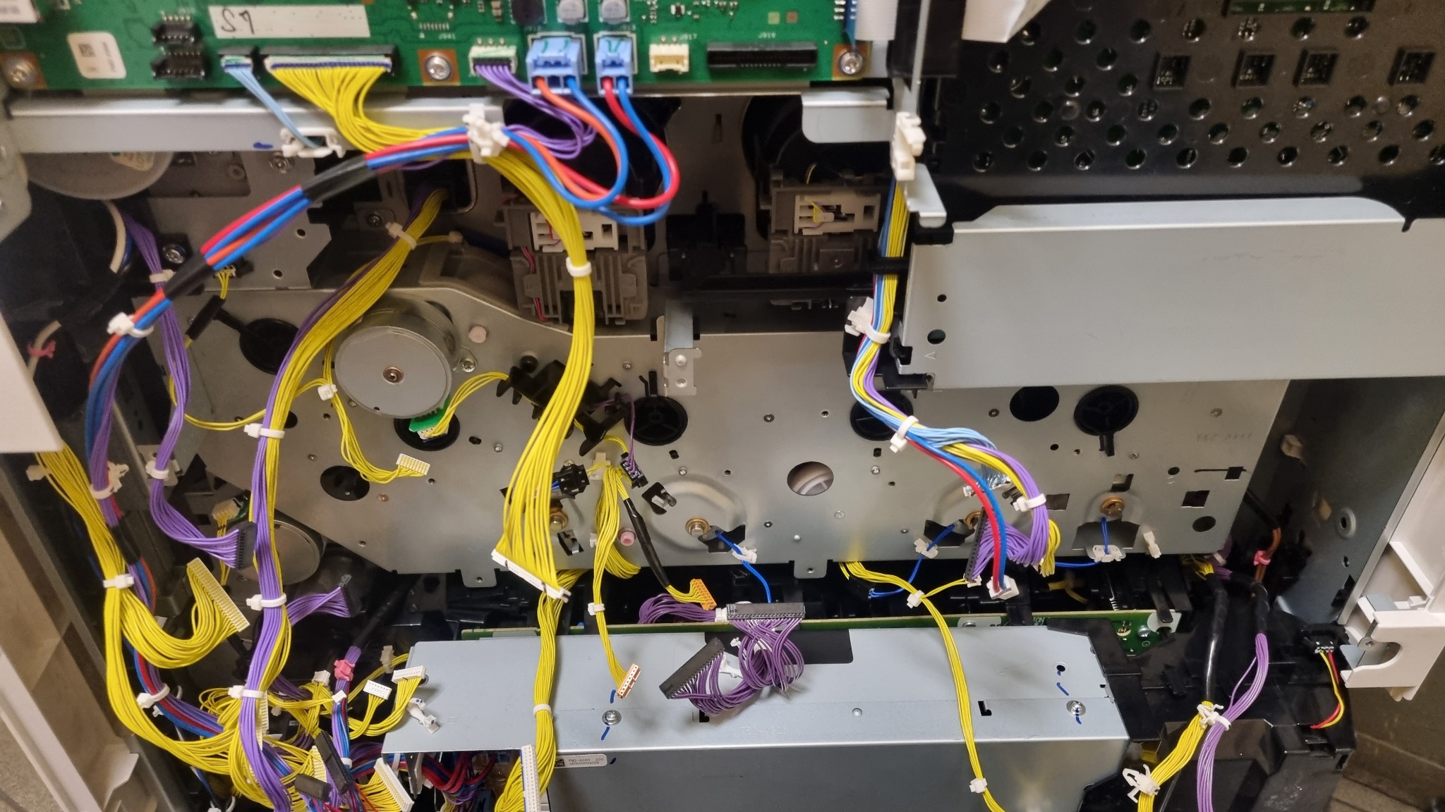 Wnętrze rozmontowanej kserokopiarki z widocznymi kablami, modułami i elementami mechanicznymi, ukazujące skomplikowaną konstrukcję urządzenia.