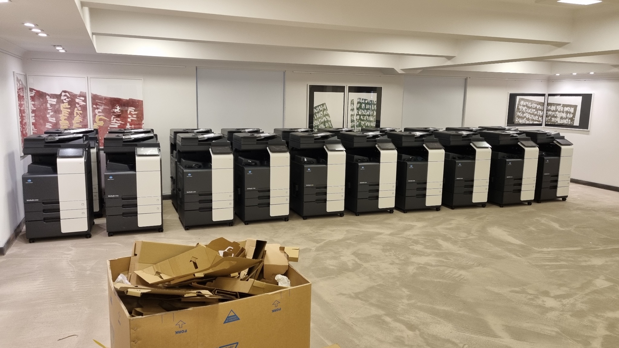 Rząd nowych, czarno-białych kserokopiarek biurowych marki Konica Minolta, ustawionych w jasnym pomieszczeniu z kartonem po rozpakowaniu na pierwszym planie.