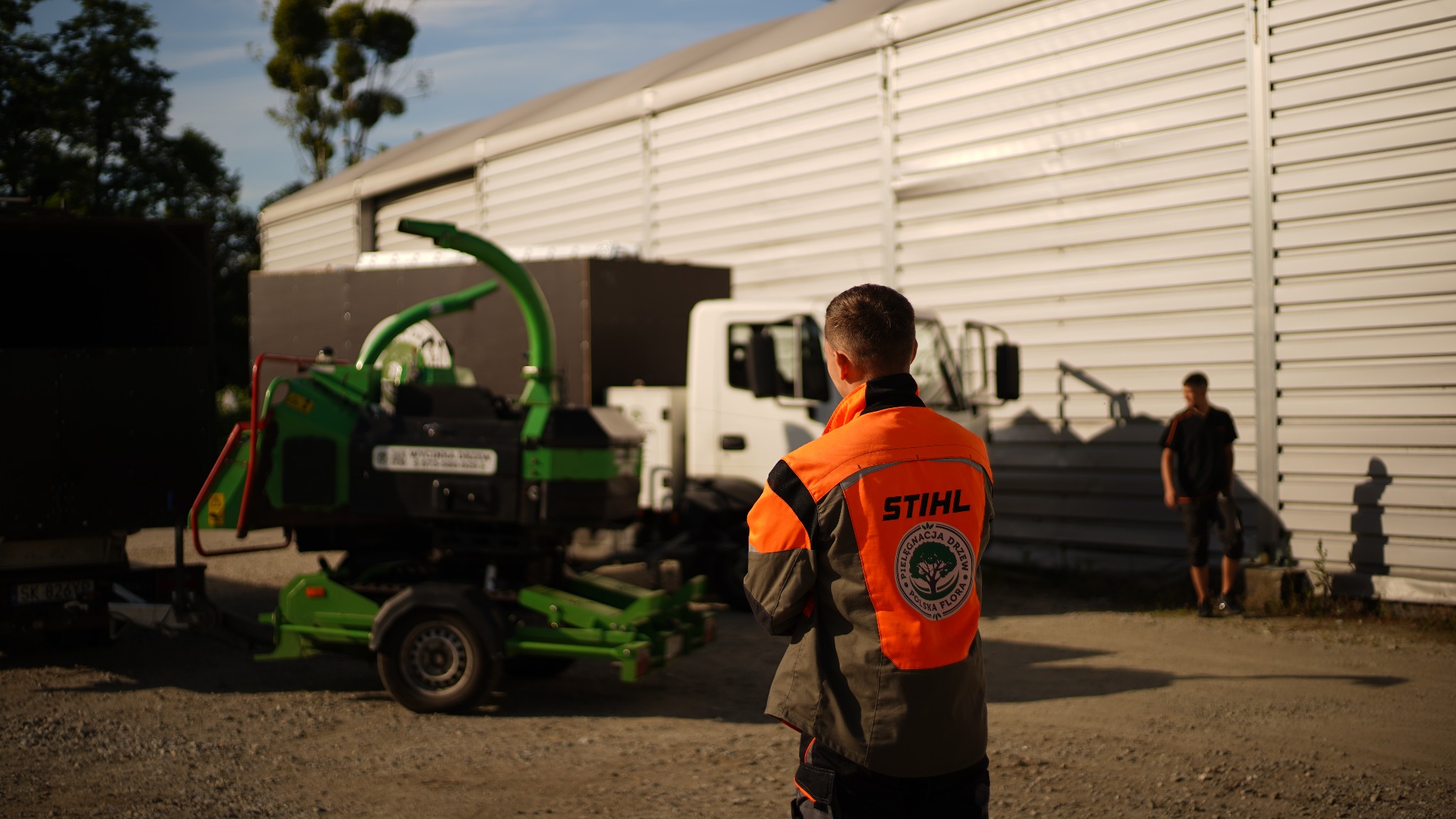 Pracownik w pomarańczowej kurtce Stihl stoi tyłem na placu budowy, obok zielonego rębaka do gałęzi i białej ciężarówki, w tle budynek z blachy falistej.