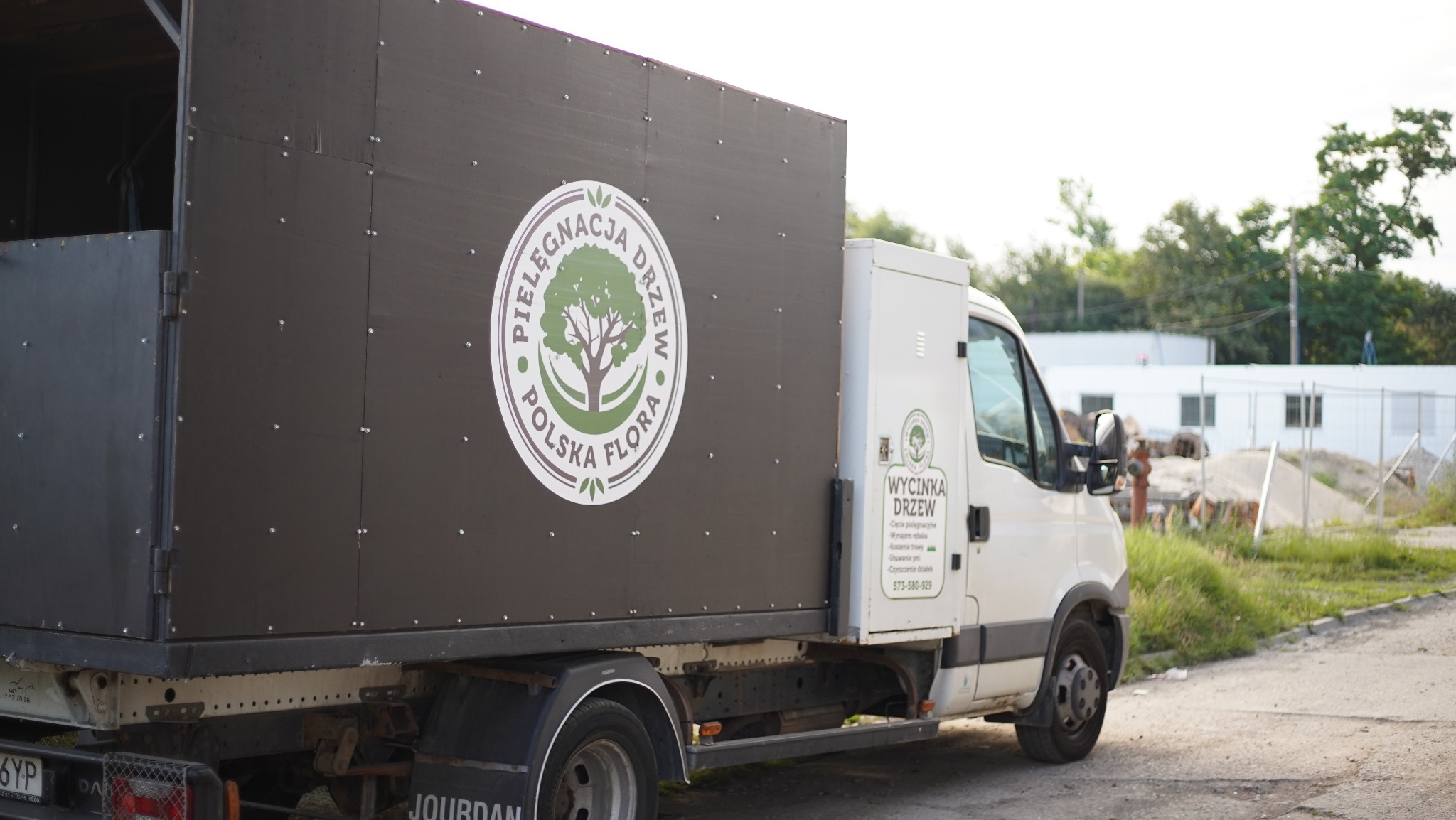 Ciężarówka firmy 'Pielęgnacja Drzew Polska Flora' z logo i danymi kontaktowymi 'Wycinka Drzew', zaparkowana na tle budynków i zieleni.