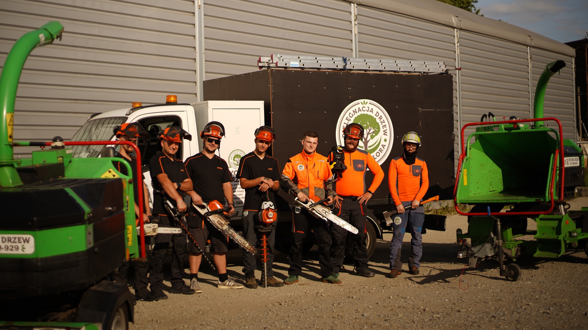 Zespół pracowników w strojach roboczych z logiem firmy, stojący przed samochodem z rozdrabniaczem gałęzi i piłą spalinową w dłoniach, gotowy do pracy.