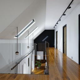 ŁUCJA ORKISZ PRIM AREA STUDIO - Nowoczesne wnętrze poddasza z drewnianą podłogą, szklaną balustradą i oknami dachowymi. Minimalistyczny design z czarnymi akcentami i wiszącymi lampami.