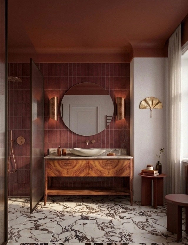 Stylowa łazienka w odcieniach bordo i bieli. Umywalka nablatowa, okrągłe lustro, marmurowa podłoga i złote akcenty tworzą elegancki wystrój. Kabina prysznicowa ze szklanymi drzwiami.