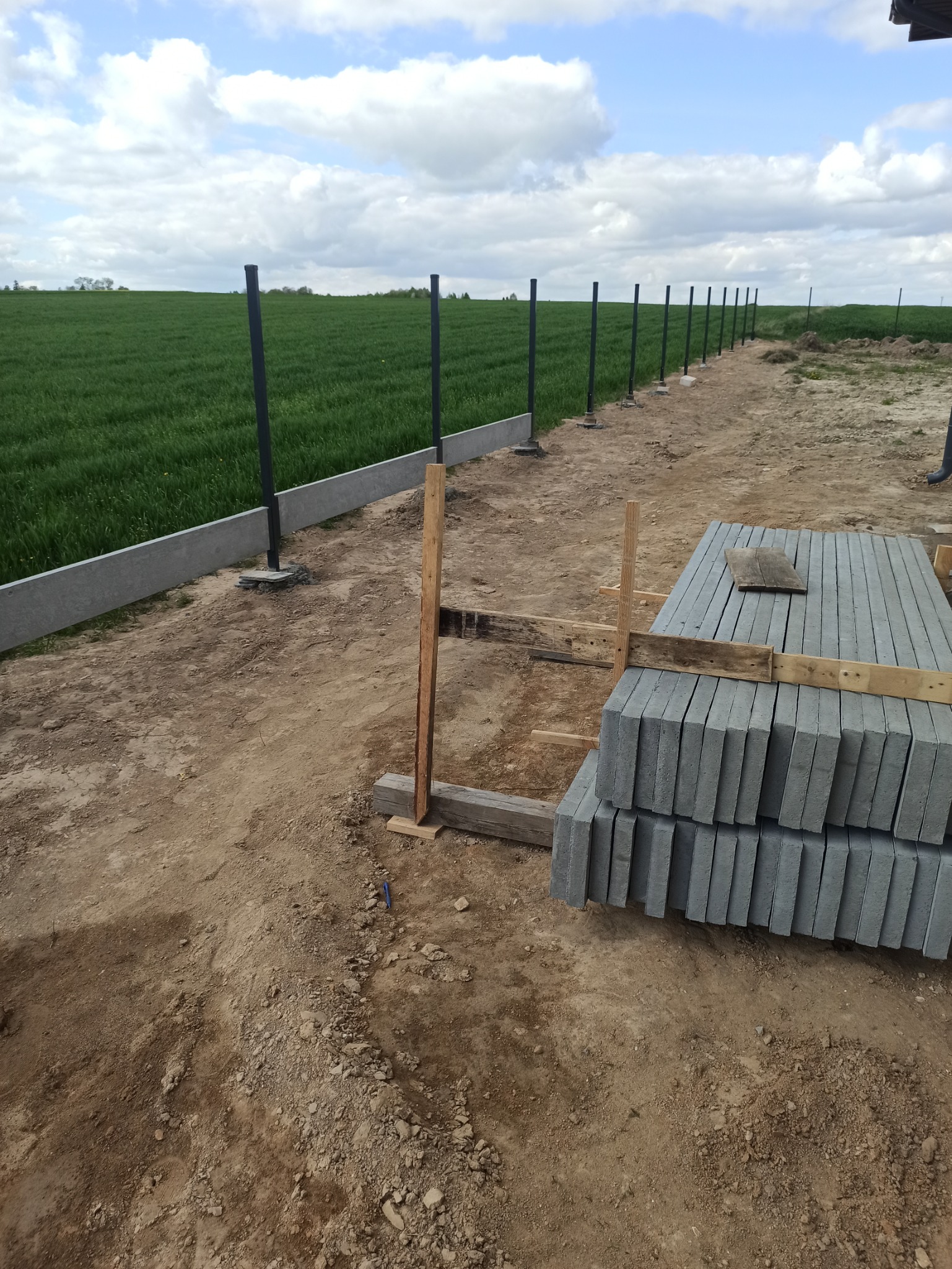 Montaż ogrodzenia panelowego na podmurówce prefabrykowanej. Widoczne słupki, betonowe płyty i stos kostki brukowej na tle zielonego pola i błękitnego nieba.