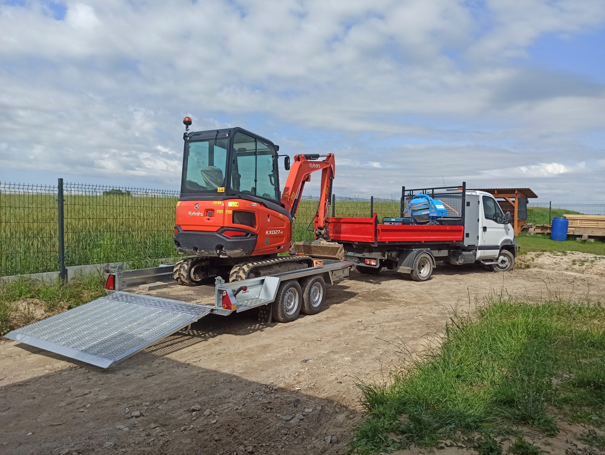 Pomarańczowa minikoparka Kubota KX027-4 na przyczepie, załadowana na ciężarówkę z czerwoną skrzynią, na tle pola i błękitnego nieba z chmurami.