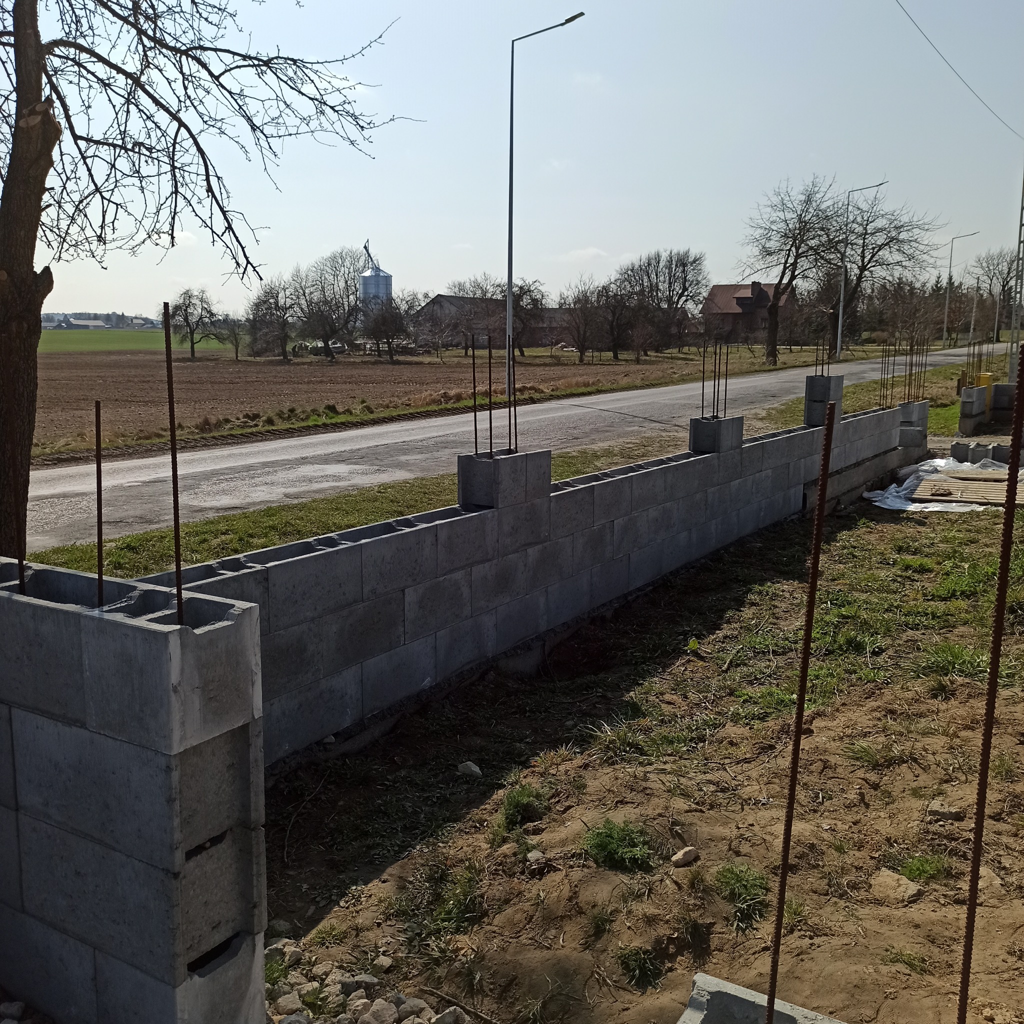 Budowa ogrodzenia z bloczków betonowych na działce z widokiem na pole i drogę. Widoczne wystające pręty zbrojeniowe i częściowo ułożone bloczki.