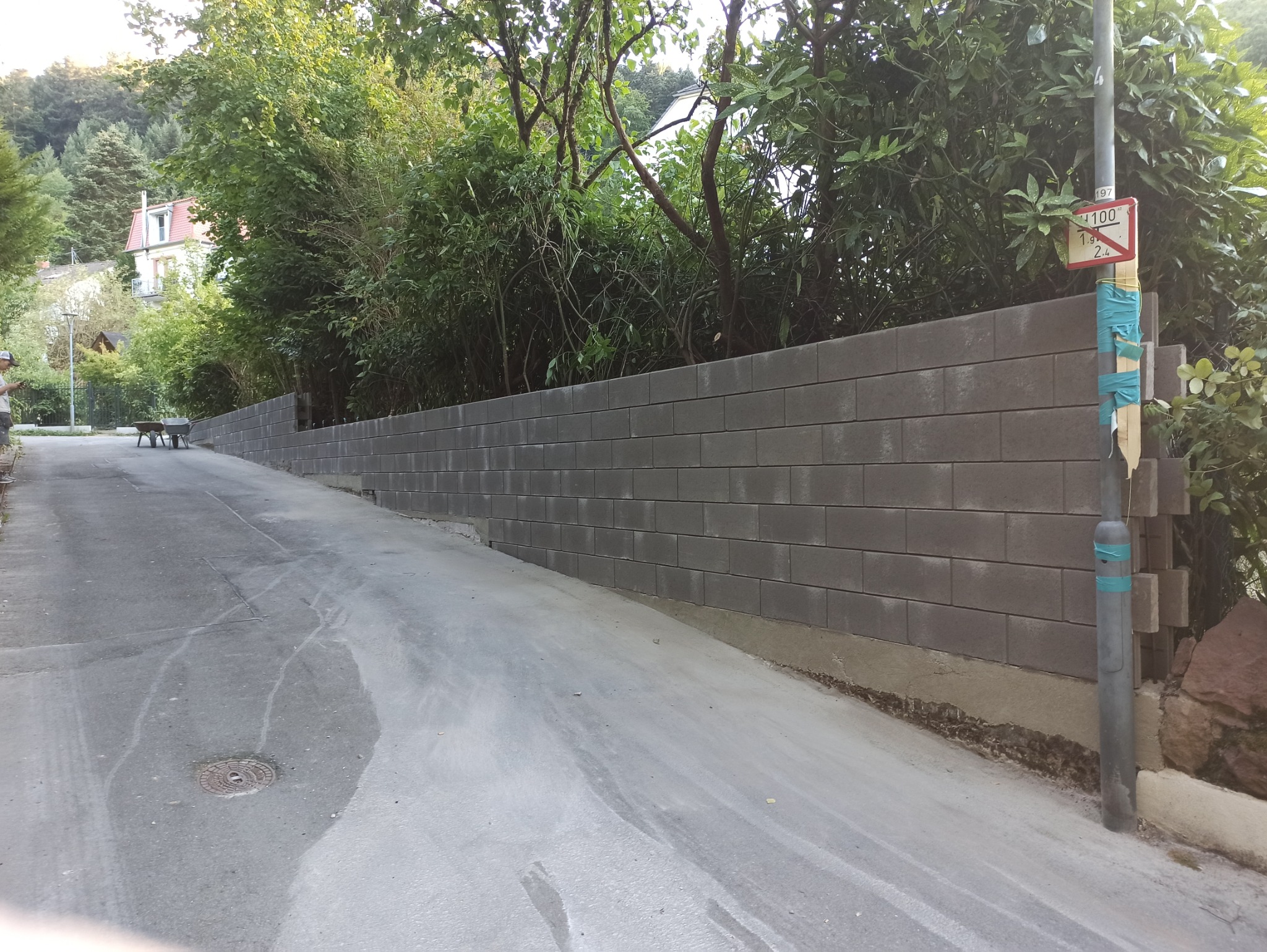 Nowo wzniesiony, szary mur z betonowych bloków na zboczu, wzdłuż asfaltowej drogi. W tle zieleń drzew i krzewów, widoczny znak drogowy na słupie.