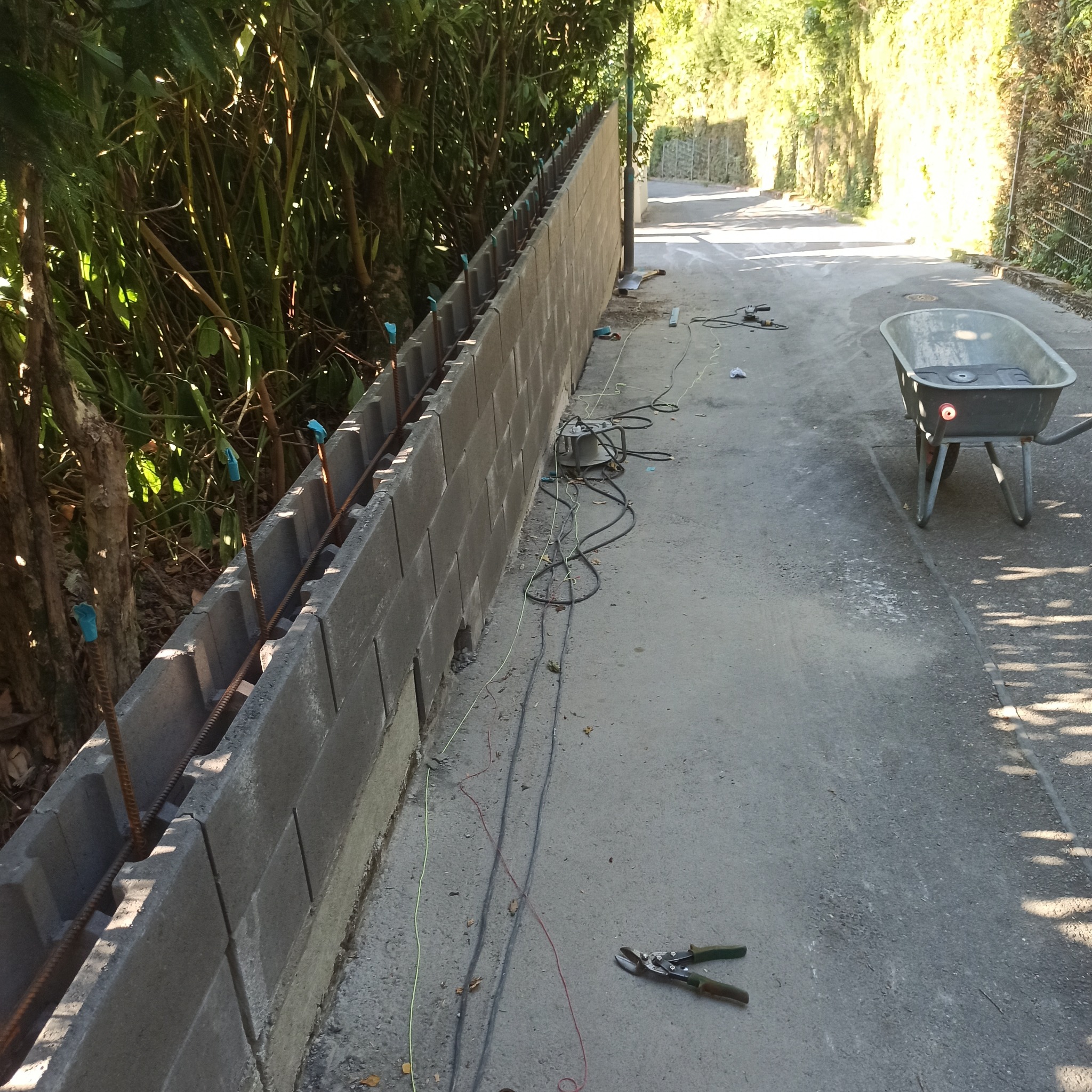 Budowa ogrodzenia z szarej bloczków betonowych, zbrojonych prętami, na tle zieleni. W pobliżu taczka i narzędzia budowlane.