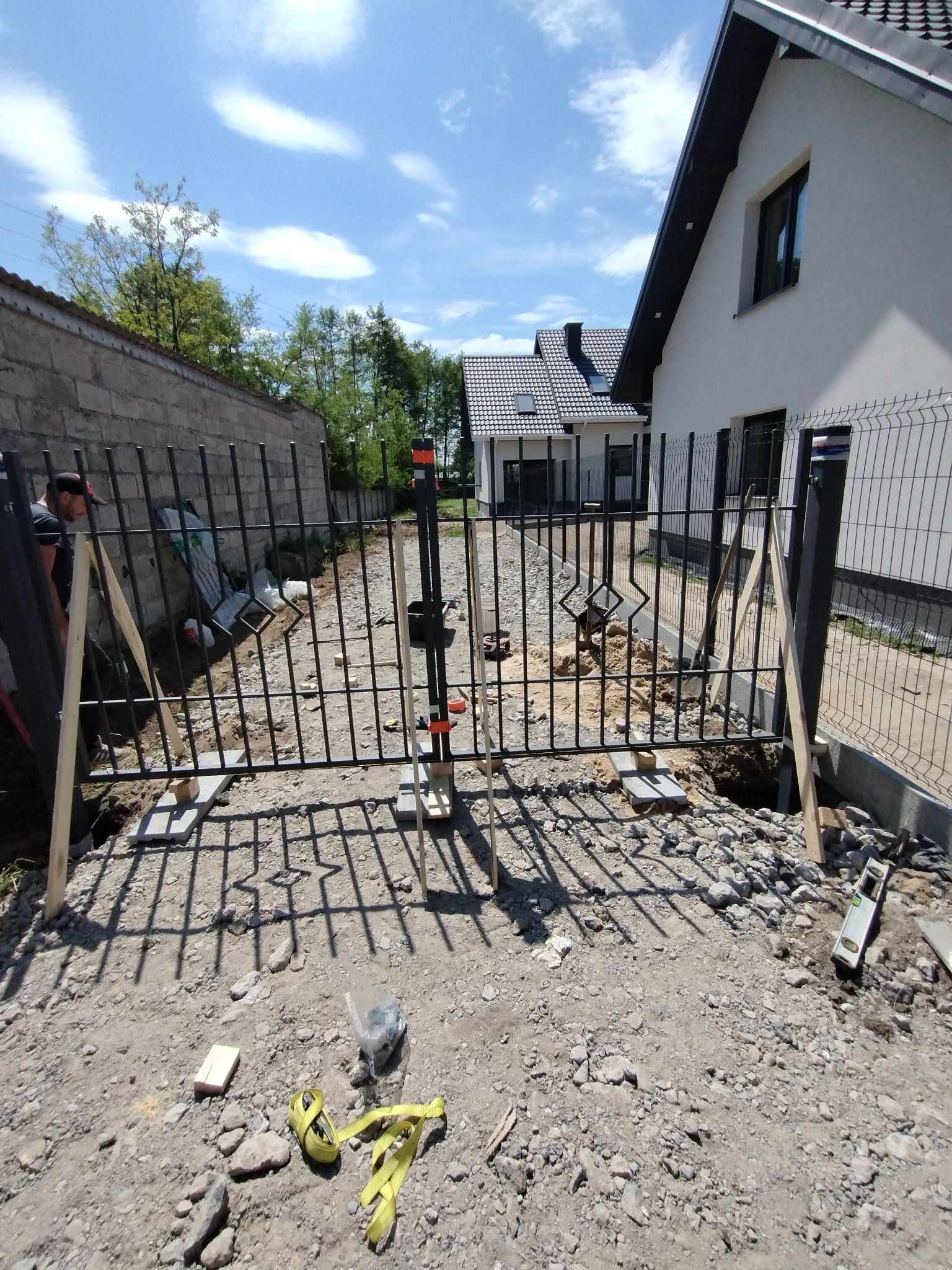 W trakcie montażu czarnej bramy ogrodzeniowej z metalowymi prętami, widoczne betonowe podstawy, poziomica, taśma miernicza i pracownik w tle, dom w nowoczesnym stylu.