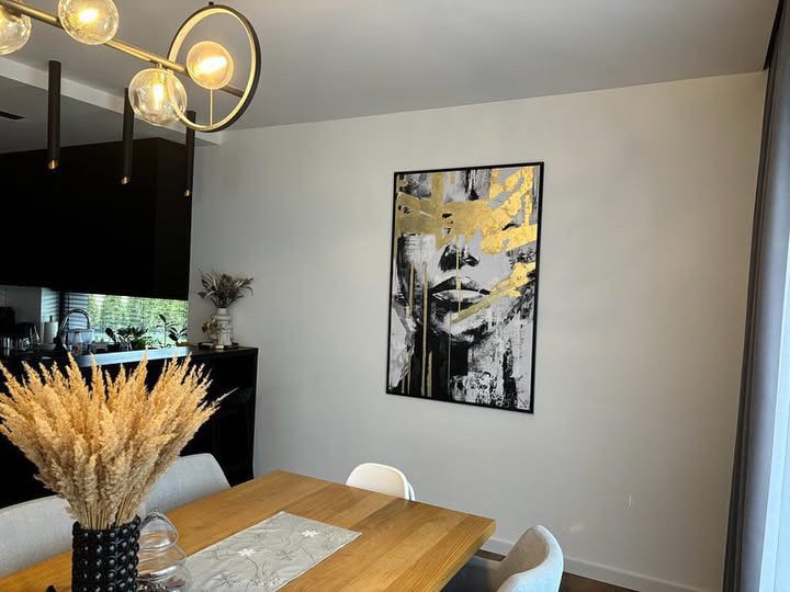 Nowoczesny salon z drewnianym stołem, bukietem suszonych traw w czarnym wazonie i abstrakcyjnym portretem kobiety z elementami złota na ścianie.