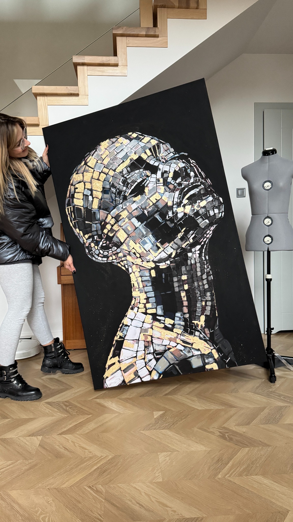 Kobieta w okularach prezentuje duży, czarny obraz z mozaikową podobizną głowy, wykonaną z lustrzanych i złotych elementów, na tle minimalistycznego wnętrza z drewnianymi schodami i manekinem...