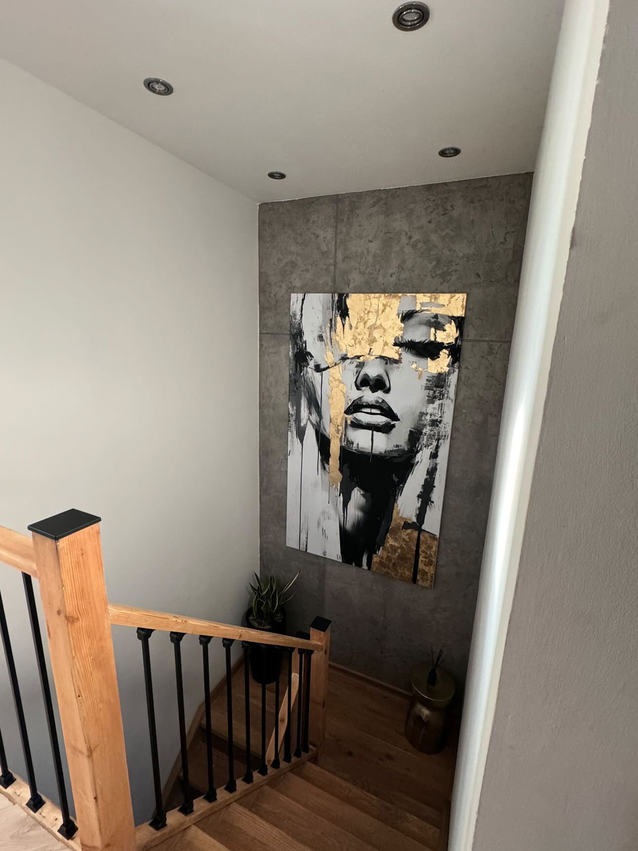 Abstrakcyjny portret kobiety z elementami złotej folii, wiszący na szarej ścianie w korytarzu obok drewnianych schodów z czarnymi balustradami.