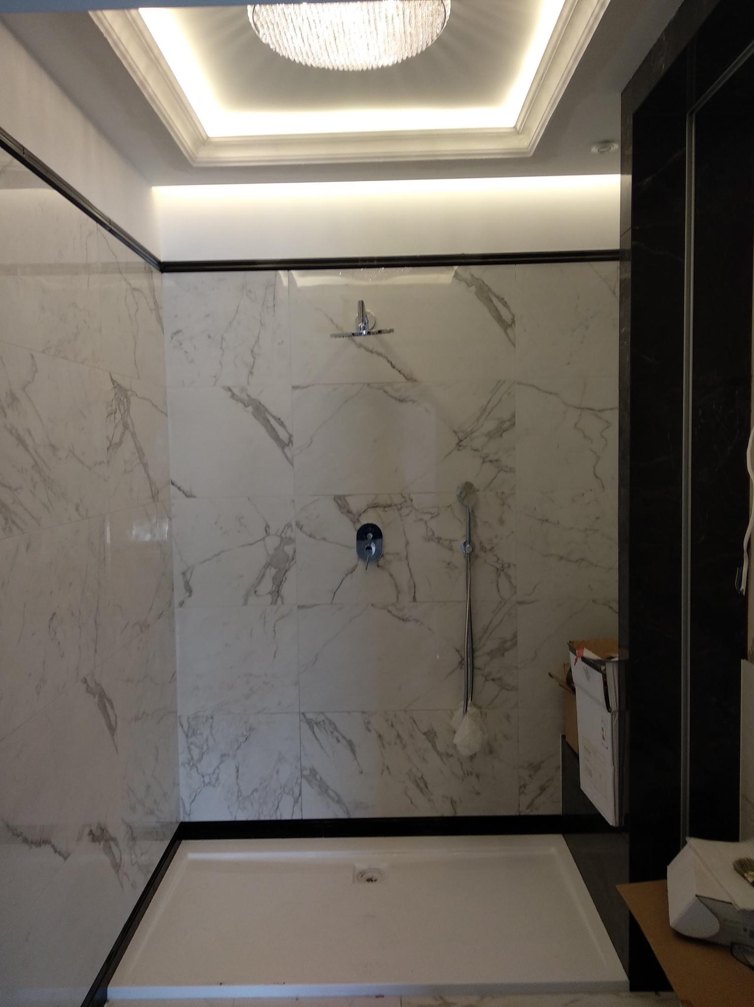 Nowoczesna kabina prysznicowa z marmurowymi płytkami, czarnymi ramami i ozdobnym oświetleniem sufitowym, widoczne elementy instalacji wodnej.