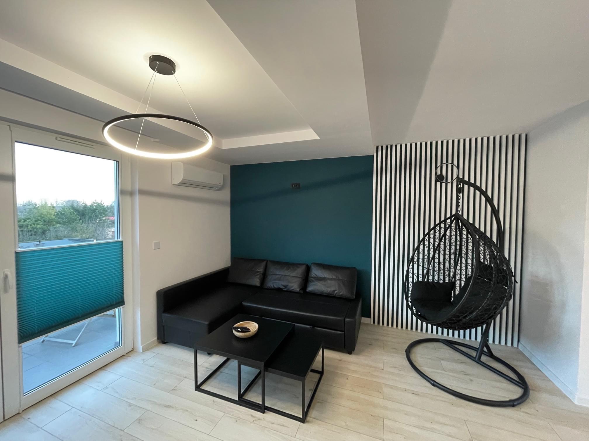 Nowoczesny salon z czarną skórzaną sofą narożną, dwoma czarnymi stolikami kawowymi, wiszącym fotelem typu kokon i okrągłą lampą LED, z akcentem turkusowej ściany i paneli dekoracyjnych.