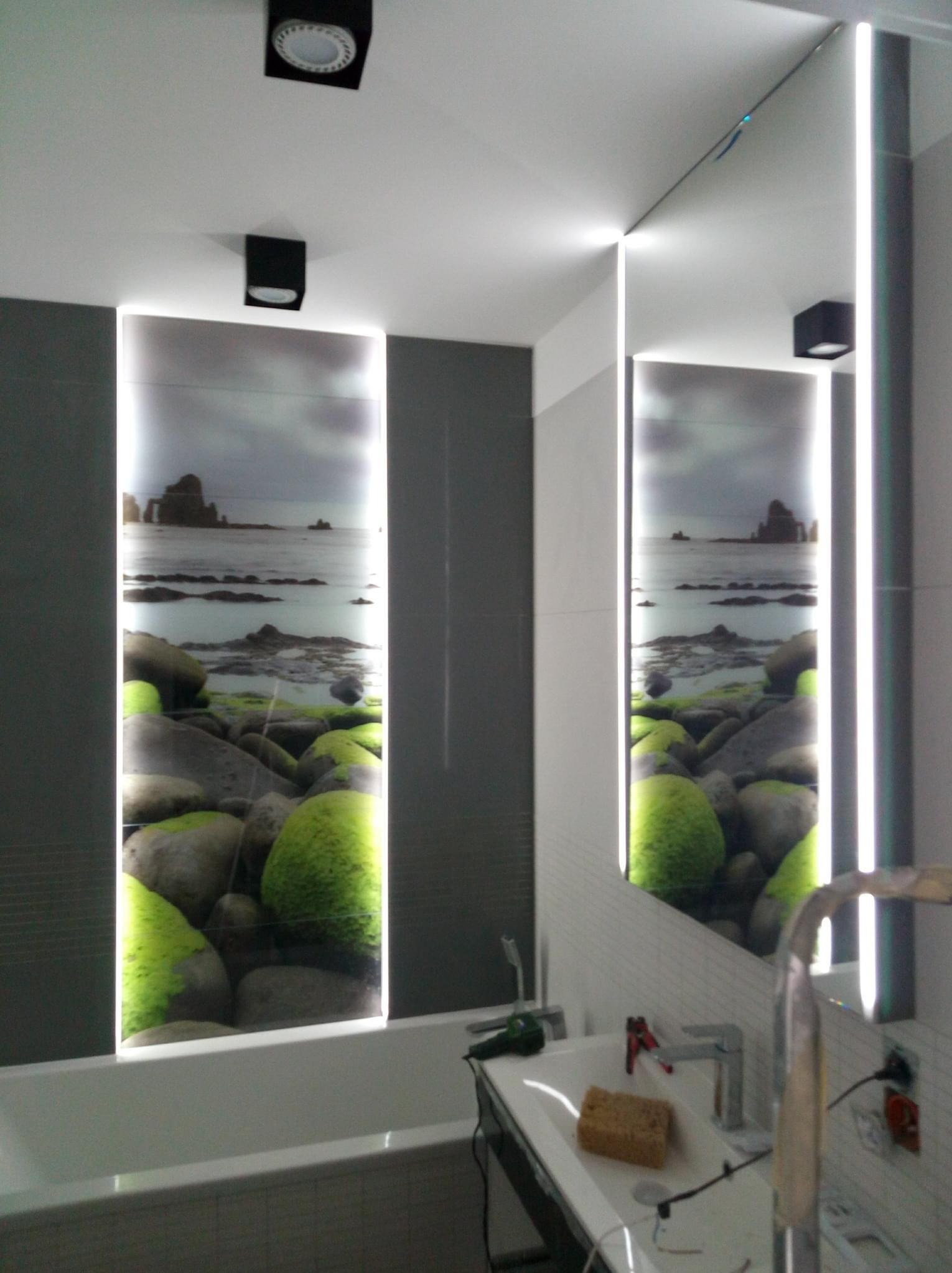 Nowoczesna łazienka z podświetlanym panelem ściennym przedstawiającym morski krajobraz z kamieniami porośniętymi mchem; widoczne elementy instalacji elektrycznej i armatura w trakcie montażu.