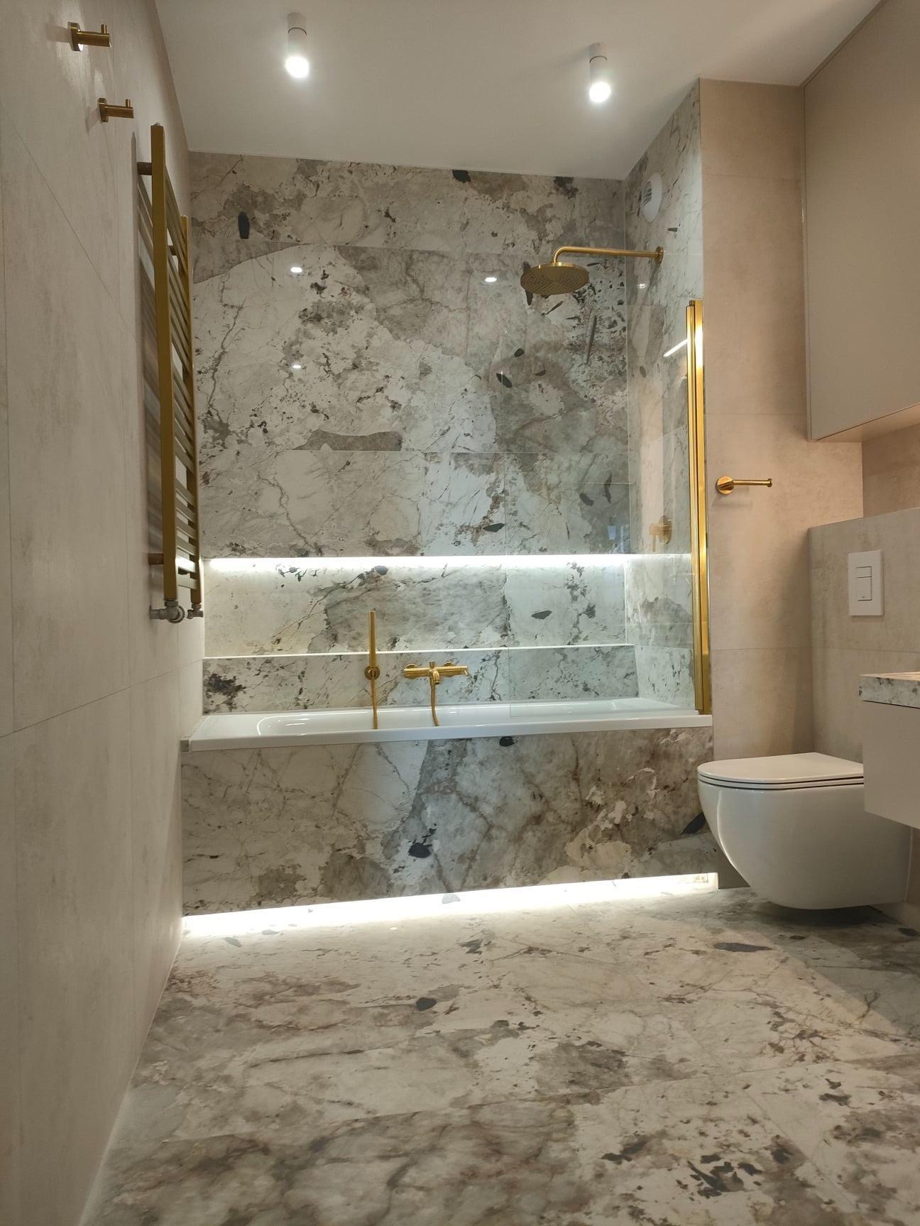 Nowoczesna łazienka z marmurowymi płytkami, złotą armaturą i podświetleniem LED pod wanną i nad nią, widok na wannę, toaletę i grzejnik ręcznikowy.