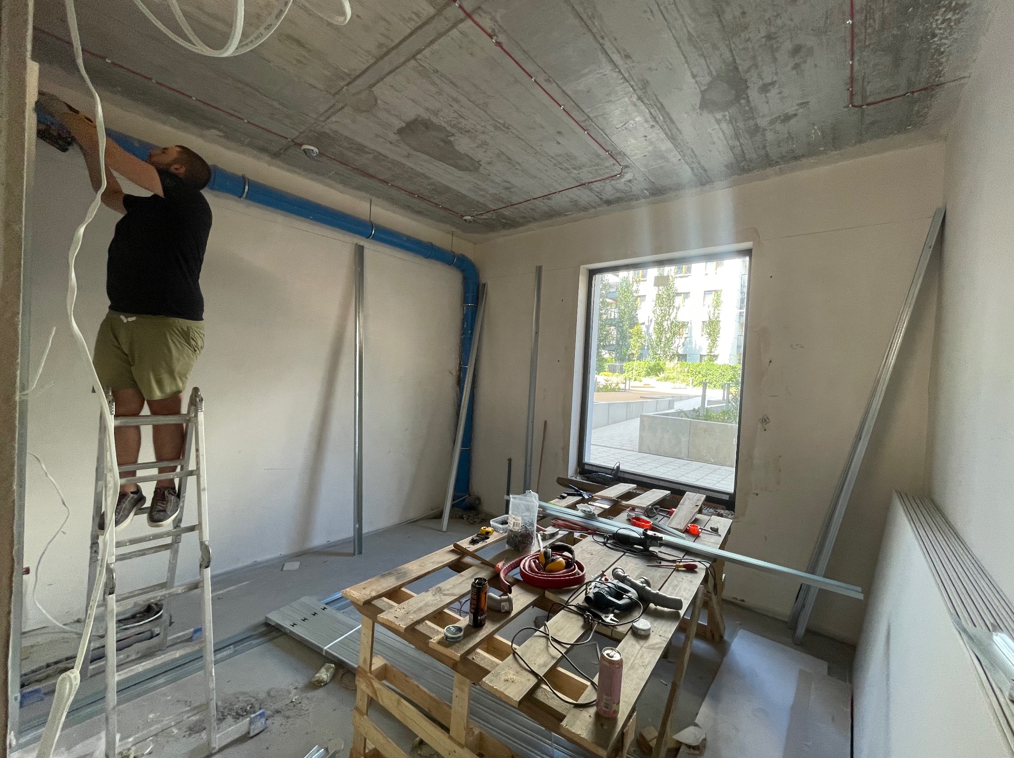 Mężczyzna na drabinie montuje płytę kartonowo-gipsową na ścianie w surowym wnętrzu z widoczną instalacją elektryczną i wentylacyjną; na stole narzędzia i materiały budowlane.