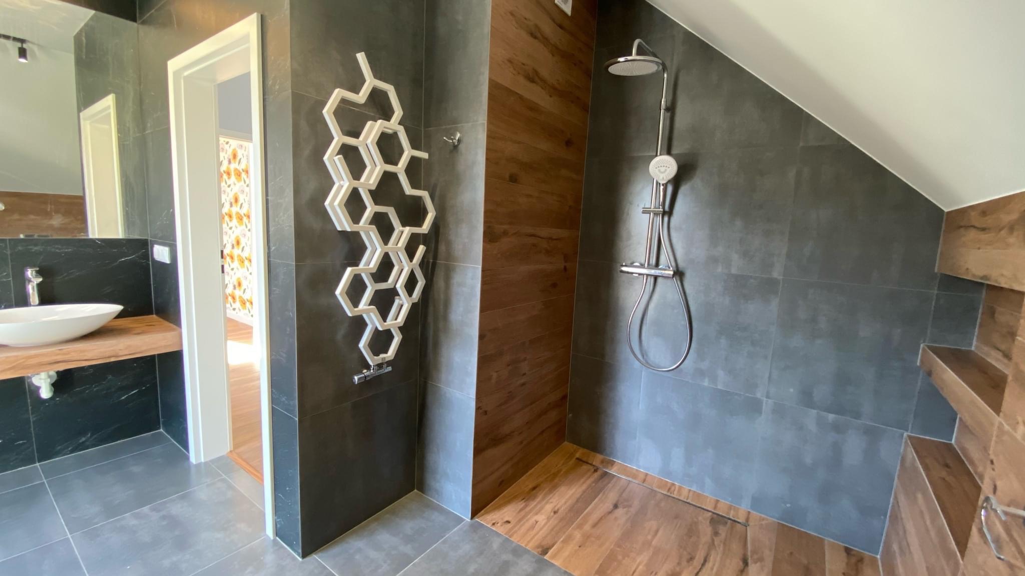 Nowoczesna łazienka z szarymi płytkami, drewnopodobnymi akcentami i grzejnikiem w kształcie plastra miodu. Widoczna strefa umywalki z blatem z naturalnego drewna oraz kabina prysznicowa typu walk-in.