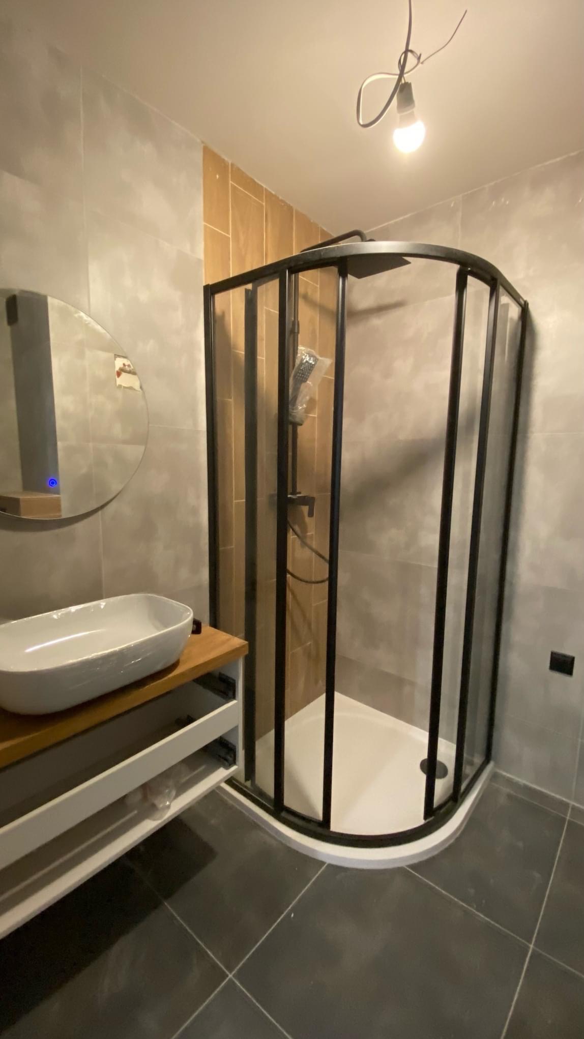 Nowoczesna łazienka z kabiną prysznicową z czarnymi ramami, umywalką nablatową na drewnianym blacie i okrągłym lustrem z podświetleniem LED, szare płytki na ścianach i czarna podłoga.