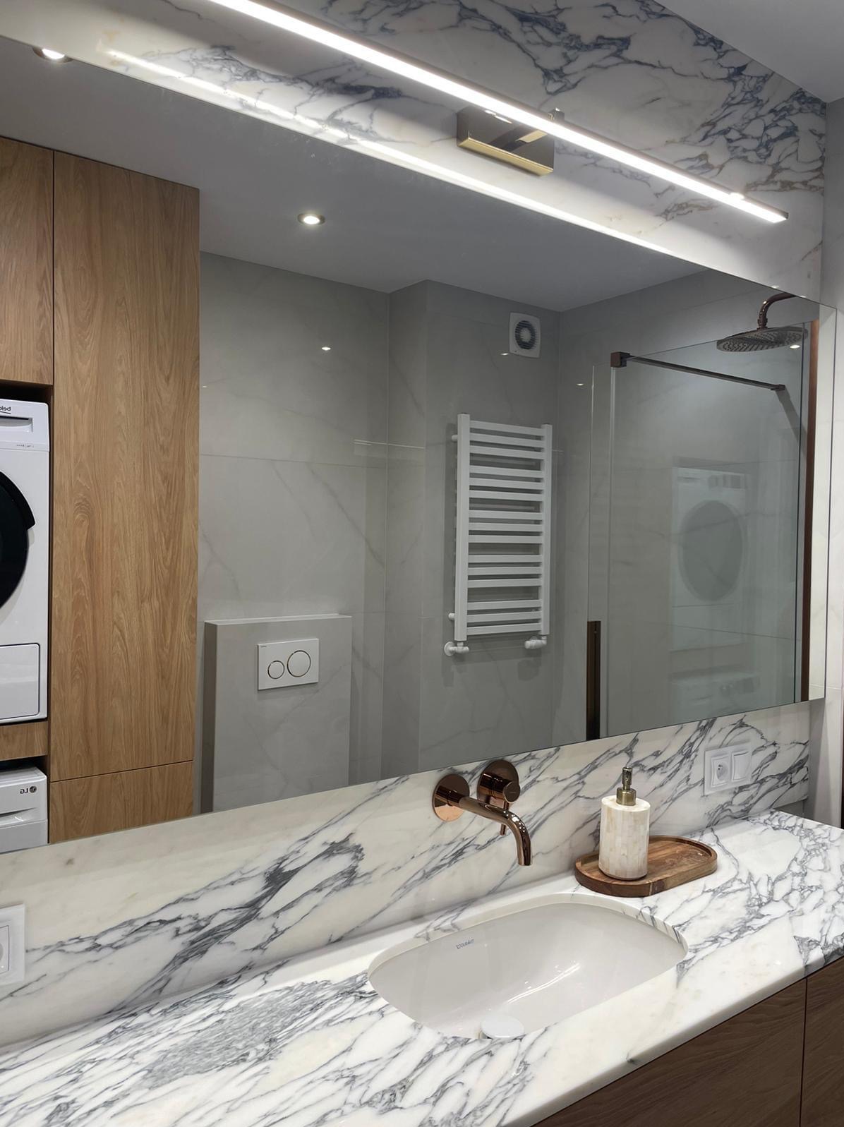 Lustro odbijające fragment łazienki z marmurowym blatem, miedzianą armaturą i grzejnikiem drabinkowym; w tle widoczny prysznic i pralka.