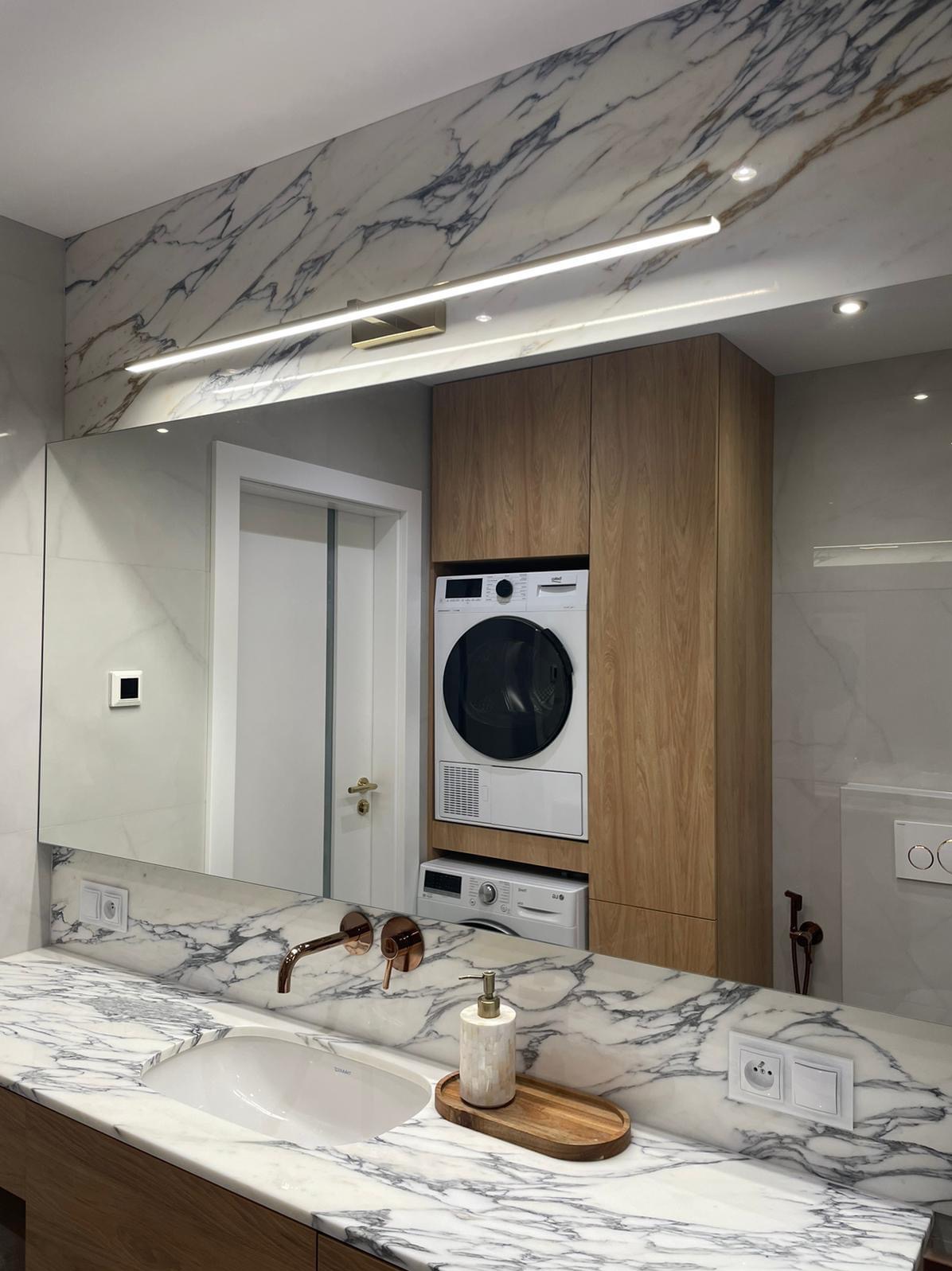 Nowoczesna łazienka z marmurowym blatem, miedzianą armaturą i pralko-suszarką w drewnianej zabudowie, oświetlona podłużną lampą LED.