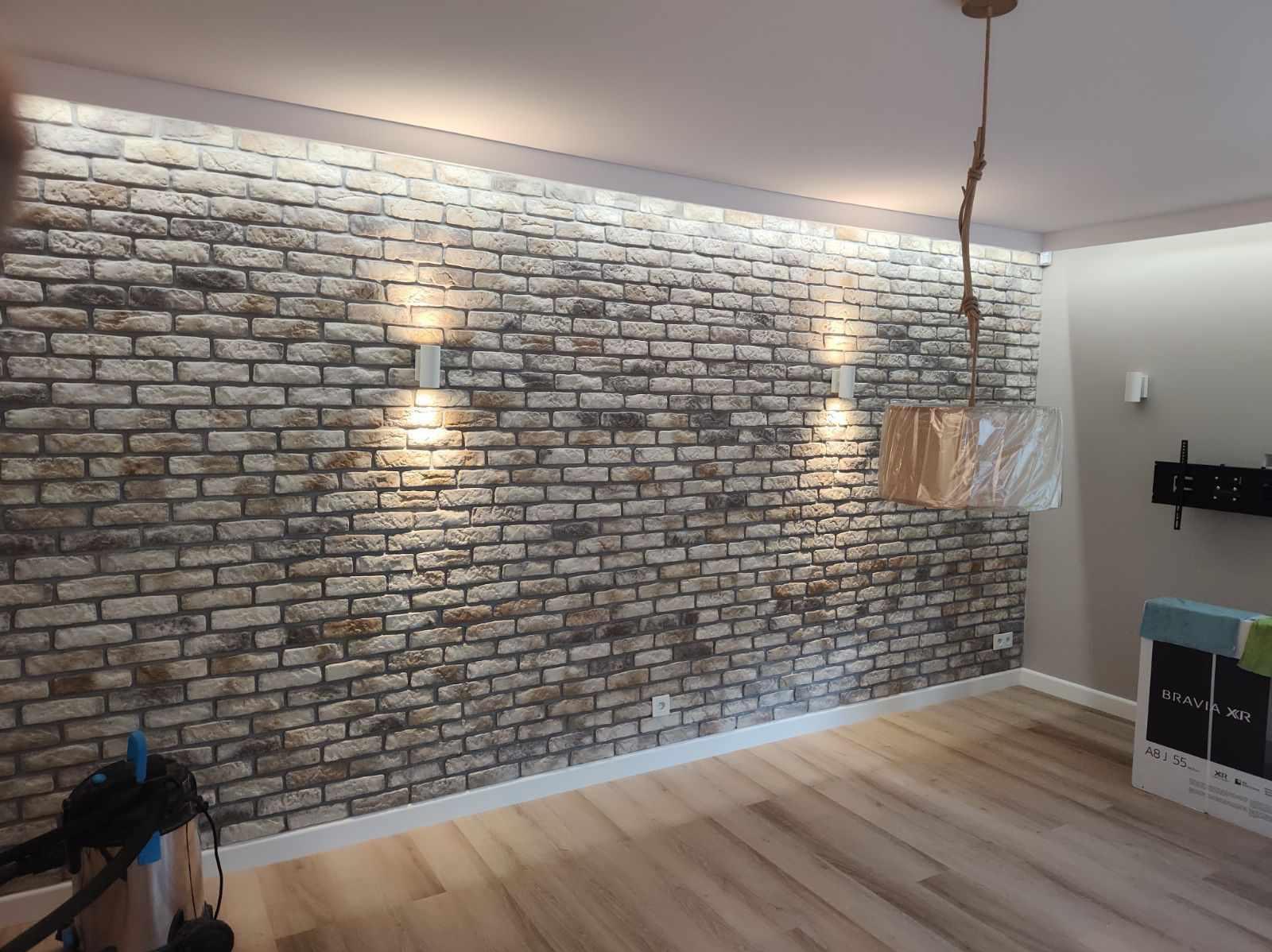 Ściana z dekoracyjnej cegły z podświetleniem LED, lampa wisząca ze sznurkowym mocowaniem, parkiet drewniany, odkurzacz i karton telewizora w rogu pokoju.