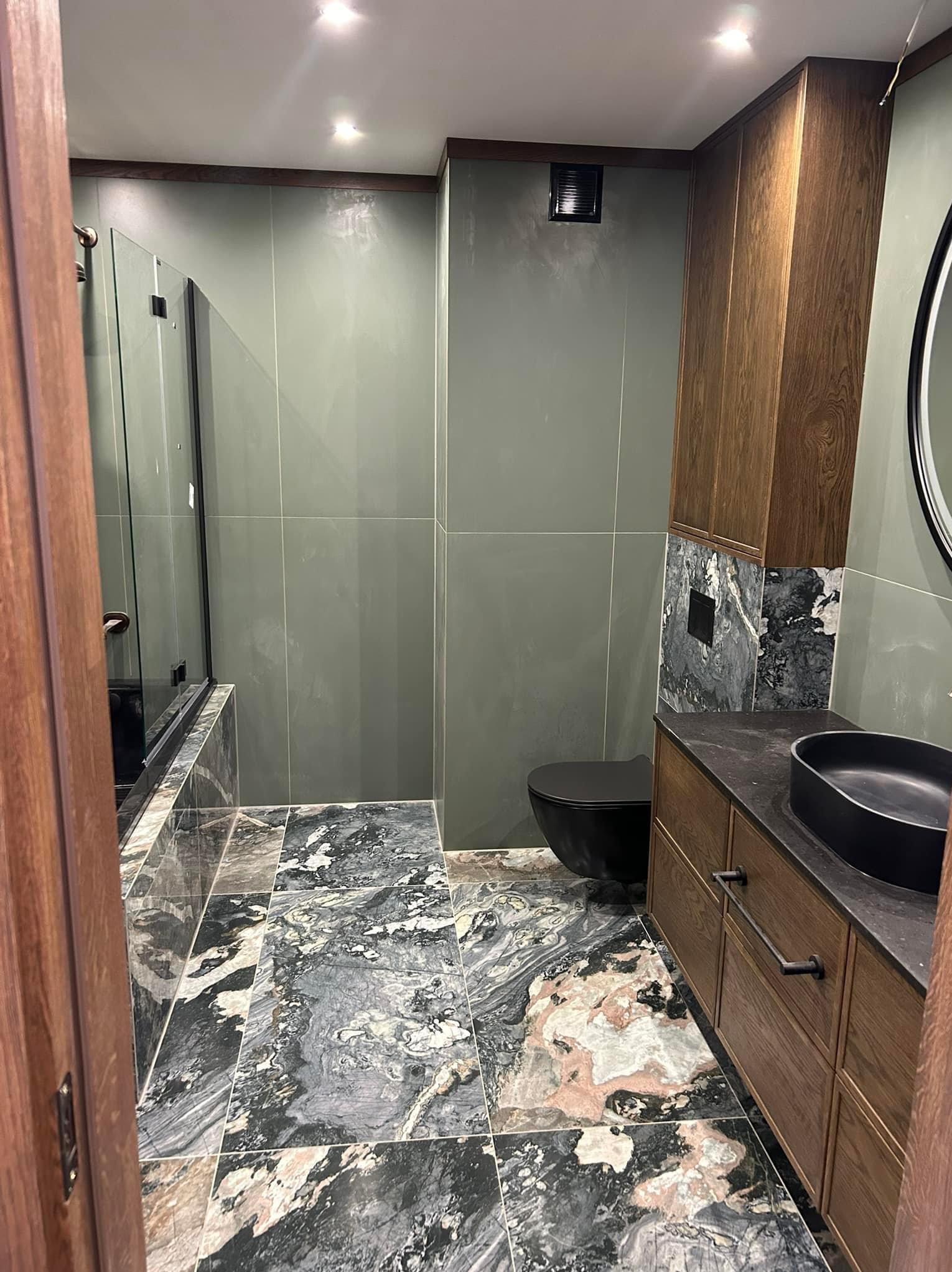 Nowoczesna łazienka z zielonymi płytkami ściennymi, marmurową podłogą, czarną armaturą i drewnianą szafką pod umywalką.