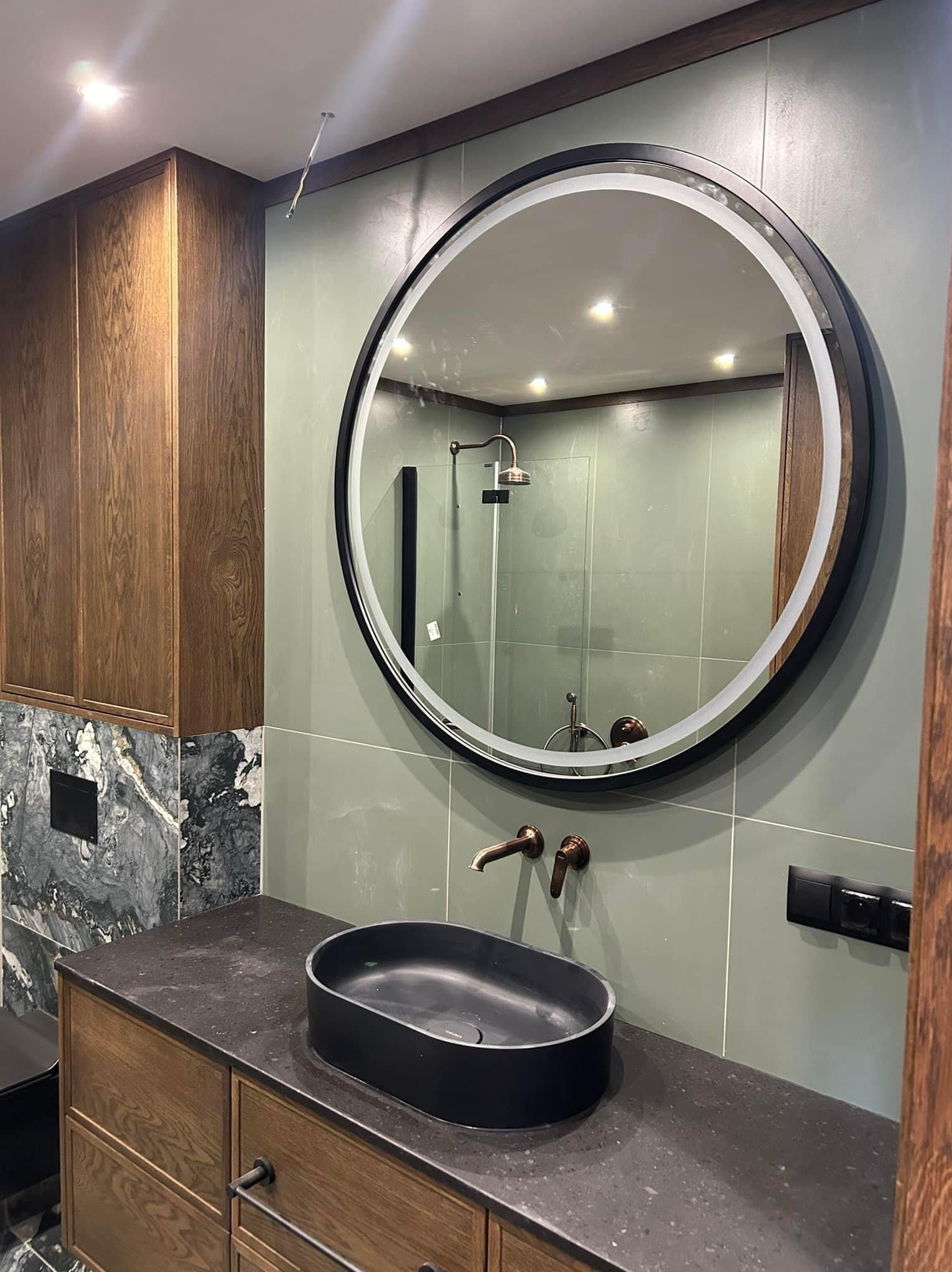 Nowoczesna łazienka z okrągłym lustrem LED, czarną umywalką nablatową i baterią w kolorze starego złota, zielone płytki na ścianach, drewniana szafka pod umywalką z marmurowym blatem.