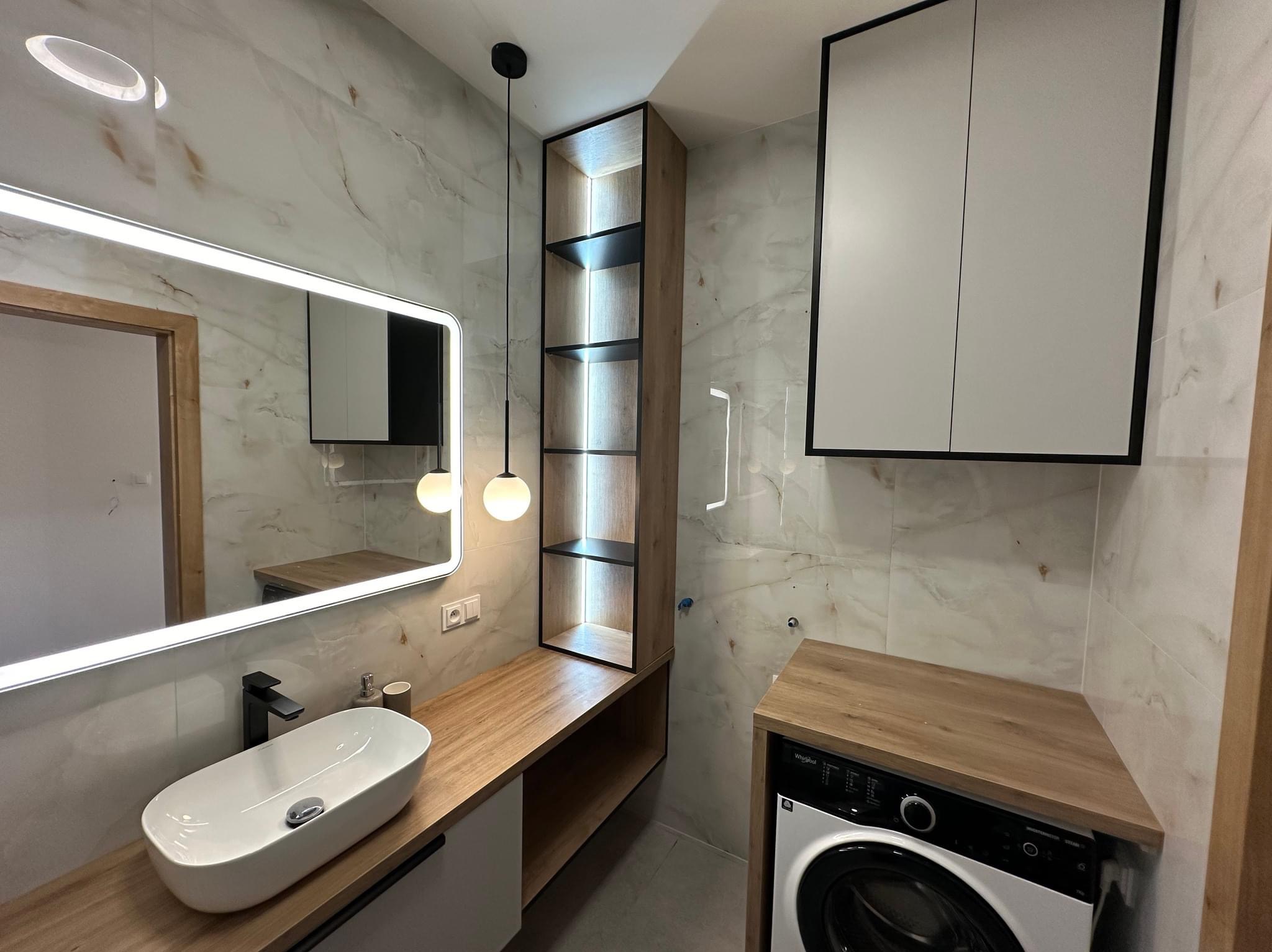 Nowoczesna łazienka z umywalką nablatową, podświetlanym lustrem, wiszącą szafką z półkami i pralką pod blatem, wykończona marmurowymi płytkami.