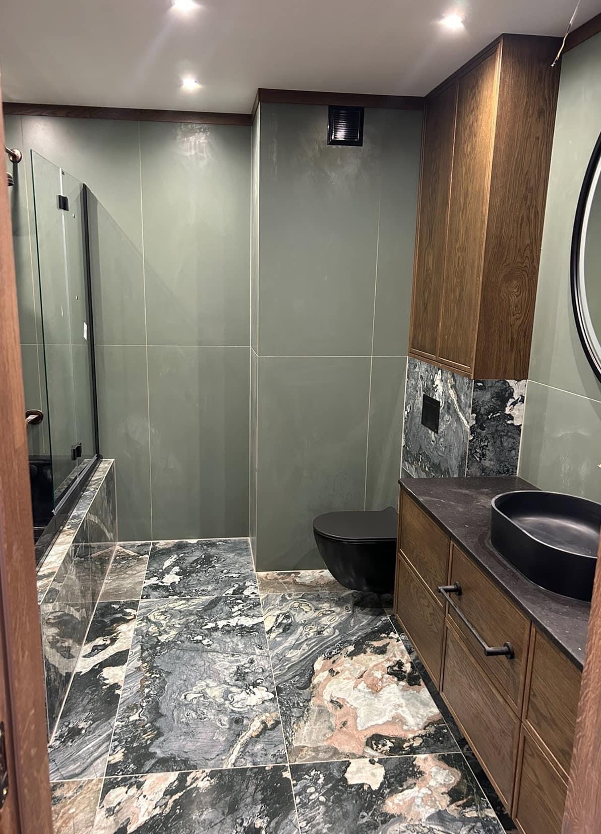 Nowoczesna łazienka z zielonymi płytkami ściennymi, marmurową podłogą, czarną armaturą i drewnianą szafką pod umywalką z marmurowym akcentem.