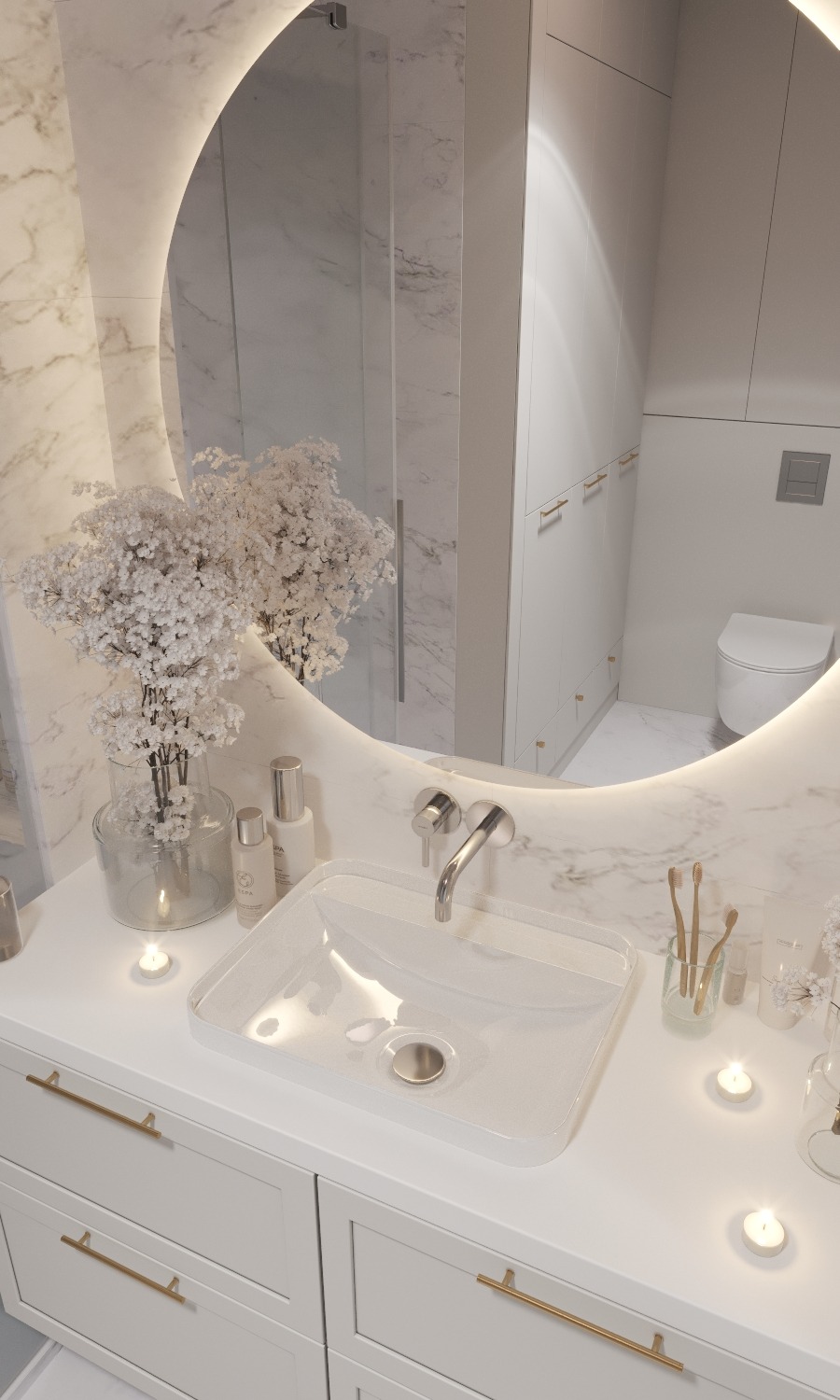 Nowoczesna łazienka z marmurowymi ścianami, okrągłym podświetlanym lustrem, szklaną umywalką i białymi szafkami z złotymi uchwytami, uzupełniona dekoracyjnymi świecami i kwiatami gipsówki.