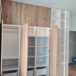 Biała szafa z drewnianymi frontami, częściowo otwarta, z oświetleniem LED wewnątrz półek i szuflad. Widoczne drążki na ubrania i wysuwane szuflady.