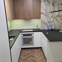 Nowoczesna kuchnia z drewnianymi szafkami górnymi, białymi szafkami dolnymi i blatem z czarnego kamienia, marmurowa ściana w tle, parkiet w jodełkę na podłodze.