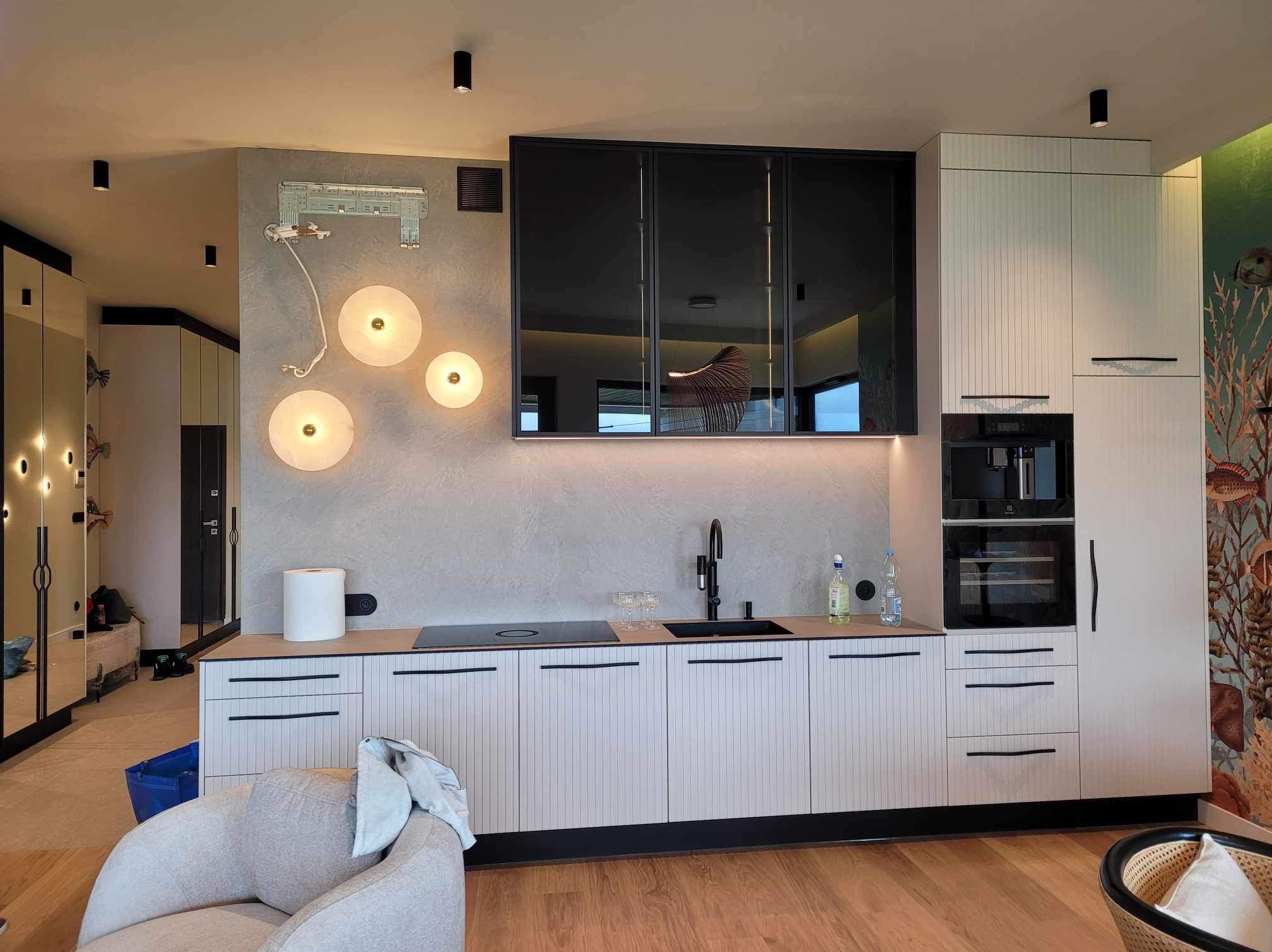 Nowoczesna kuchnia z białymi szafkami z pionowymi paskami, czarnymi uchwytami i czarnymi szklanymi szafkami wiszącymi; na ścianie dekoracyjne, okrągłe lampy i widoczny fragment instalacji; podłoga...