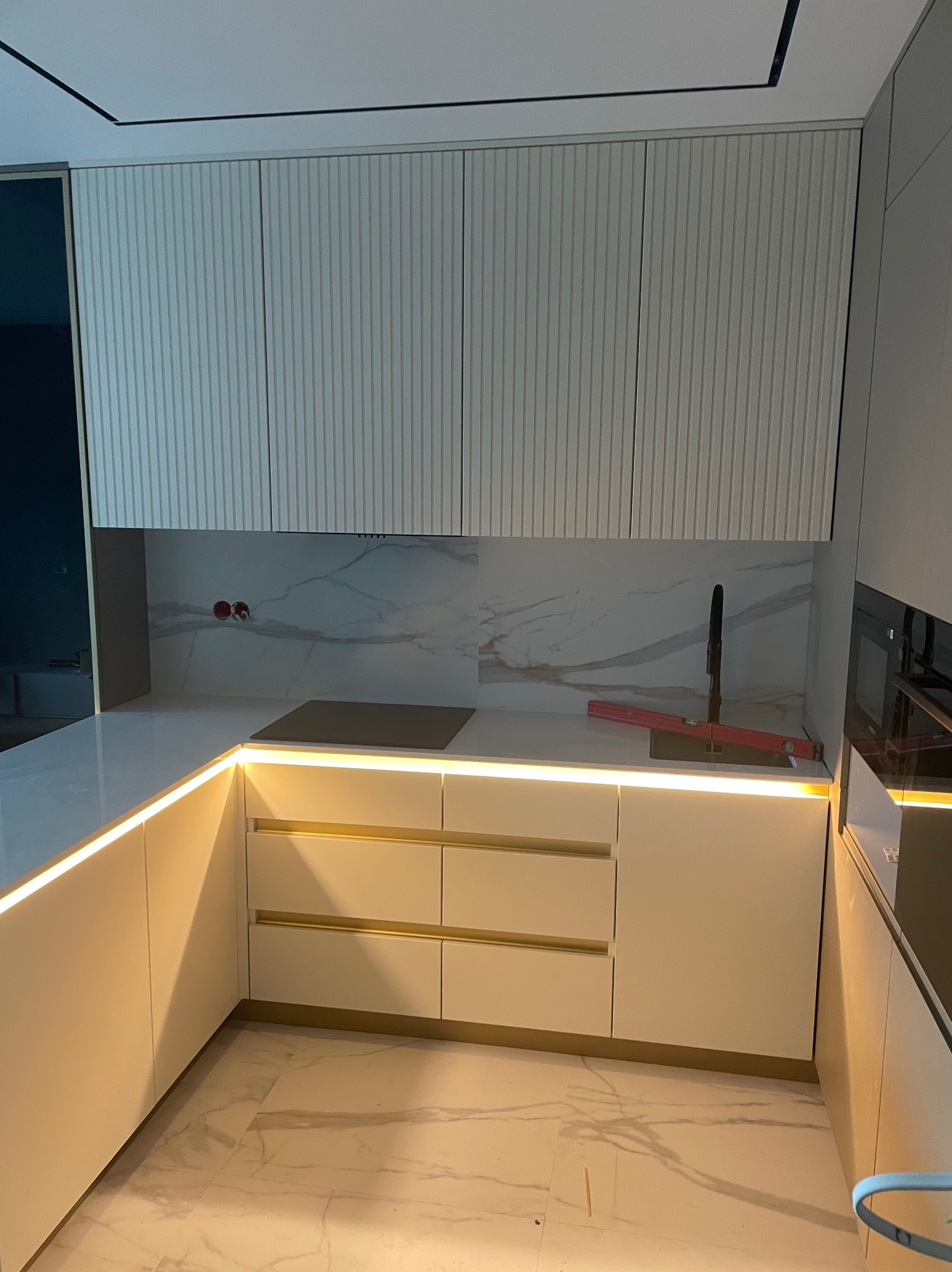 Nowoczesna kuchnia z białymi, ryflowanymi szafkami górnymi, marmurowym blatem i podświetleniem LED pod blatem, widoczny poziomica przy zlewie.