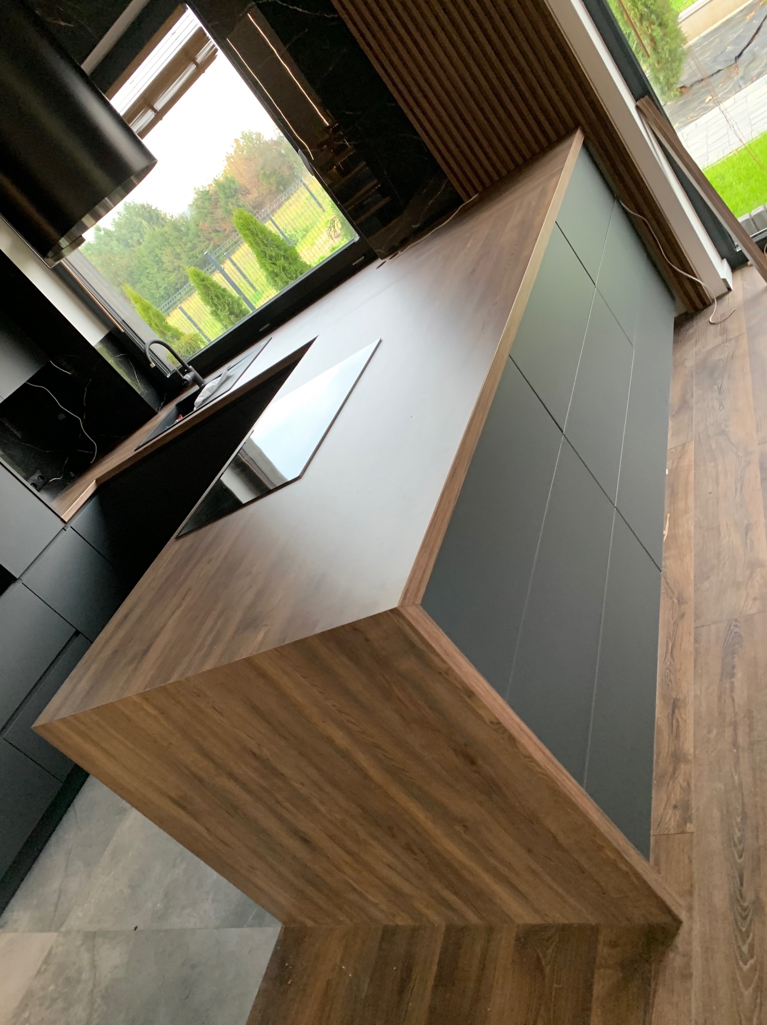 Nowoczesna kuchnia z czarnymi szafkami i drewnianym blatem, widok z góry na płytę indukcyjną i zlew przy oknie.