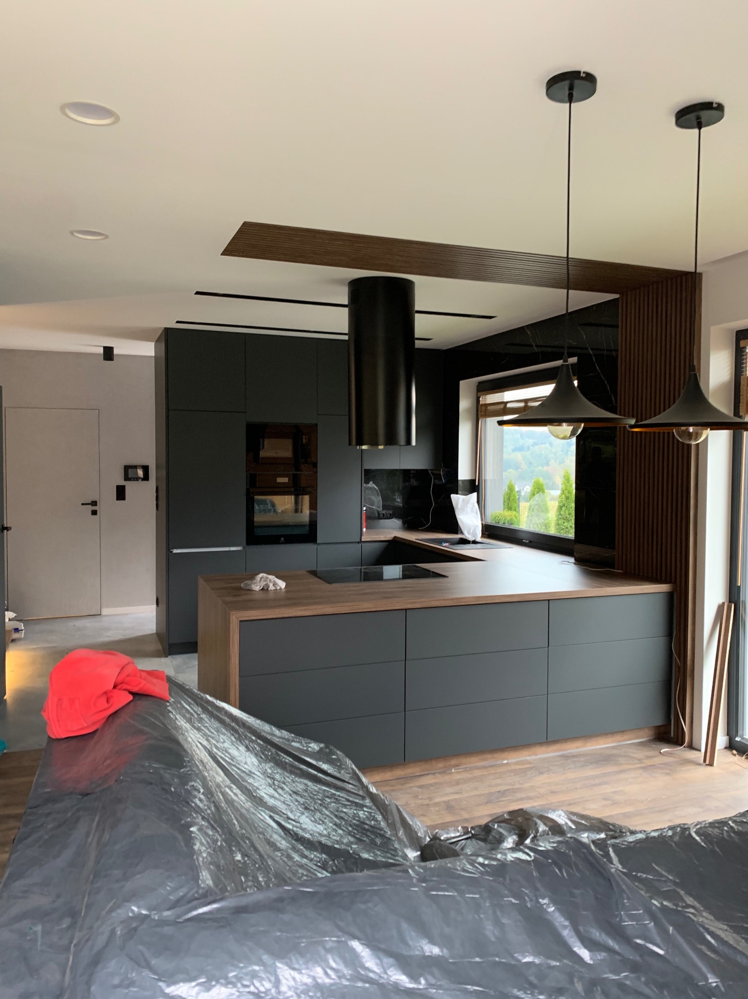 Nowoczesna kuchnia z czarnymi szafkami i drewnianym blatem, wyspą kuchenną z płytą indukcyjną, czarnym okapem cylindrycznym, oświetleniem wiszącym i elementami dekoracyjnymi z drewnianych listew...